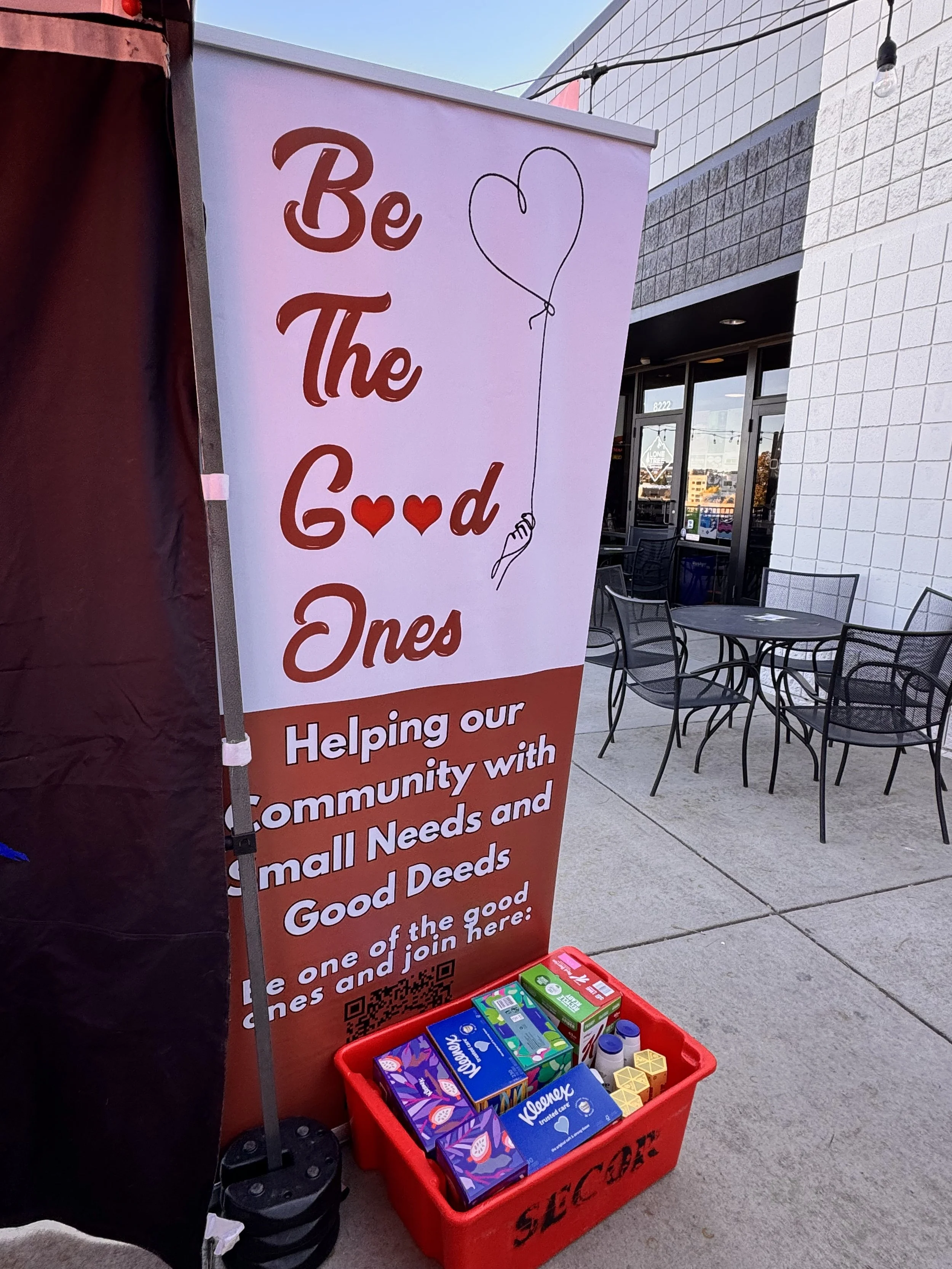 A red plastic donation bin filled with hygiene and household items, placed in front of a sign that says 'Be The Good Ones' with a heart-shaped balloon illustration and text about helping the community with small needs and good deeds.