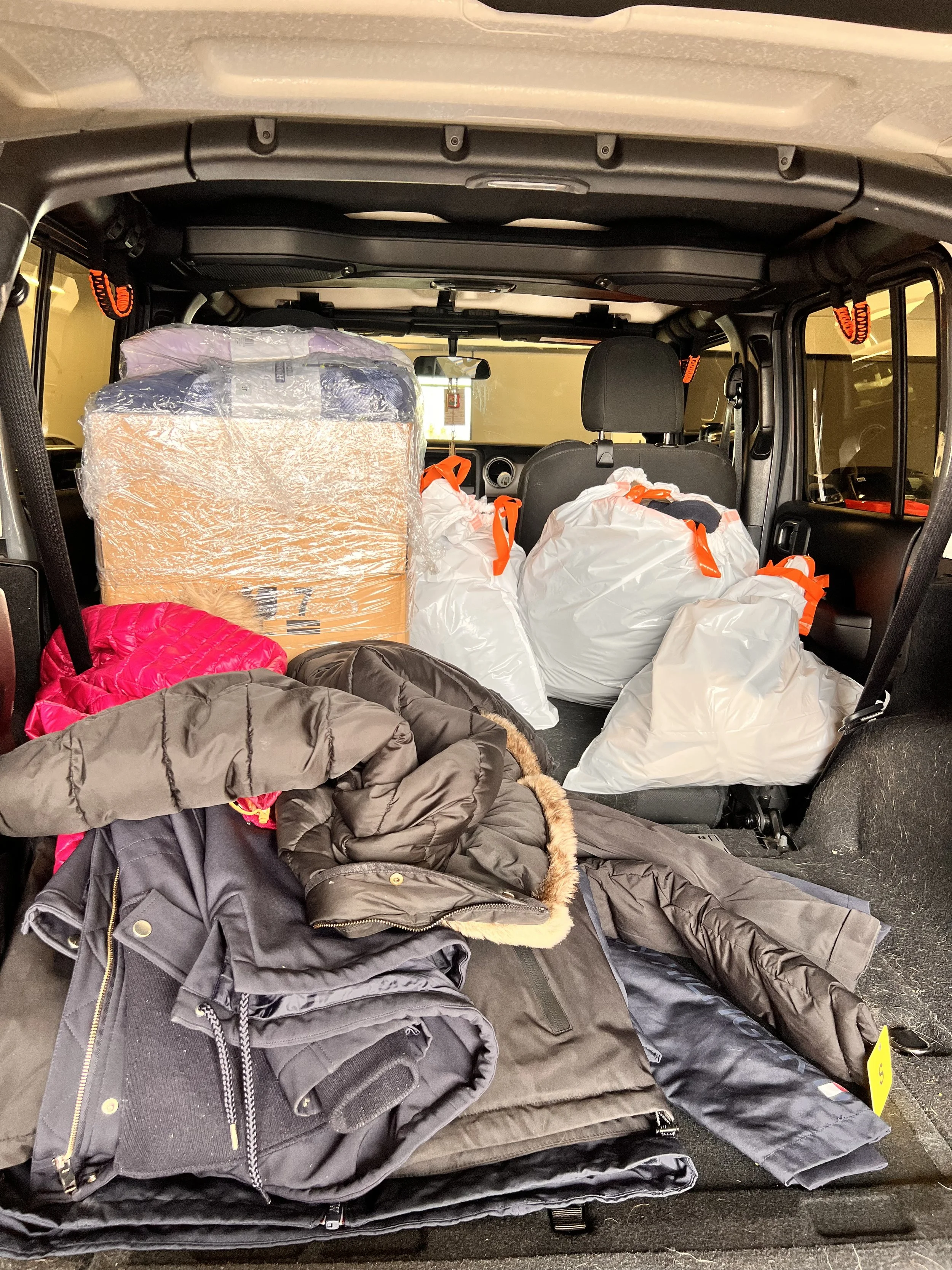 The back of a vehicle packed with piles of jackets, bags, a large cardboard box, and white plastic bags with orange handles.