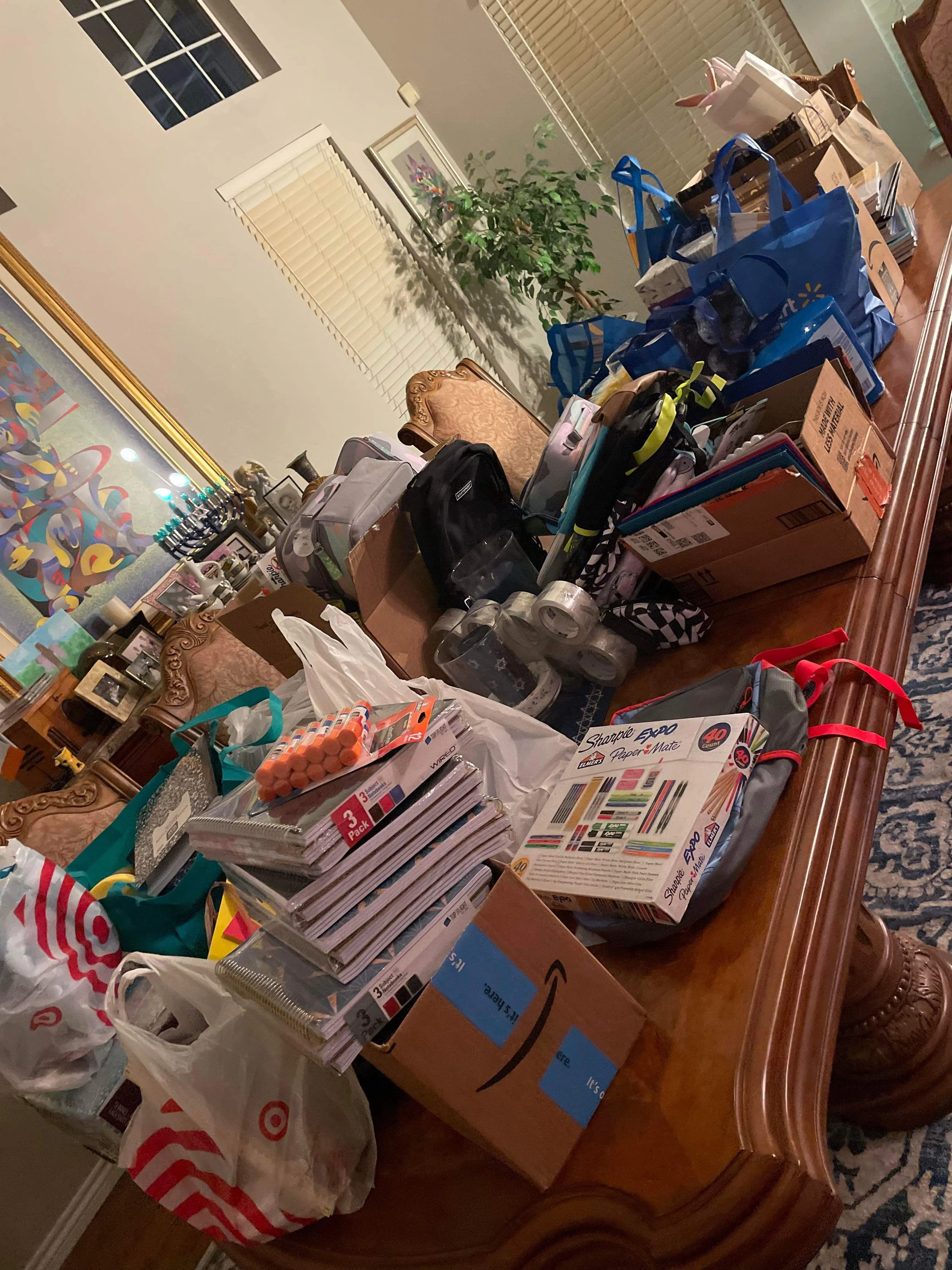 A dining room table filled with various shopping bags, boxes, and school supplies, including notebooks, markers, and tape, with chairs around it and a large potted plant in the background.
