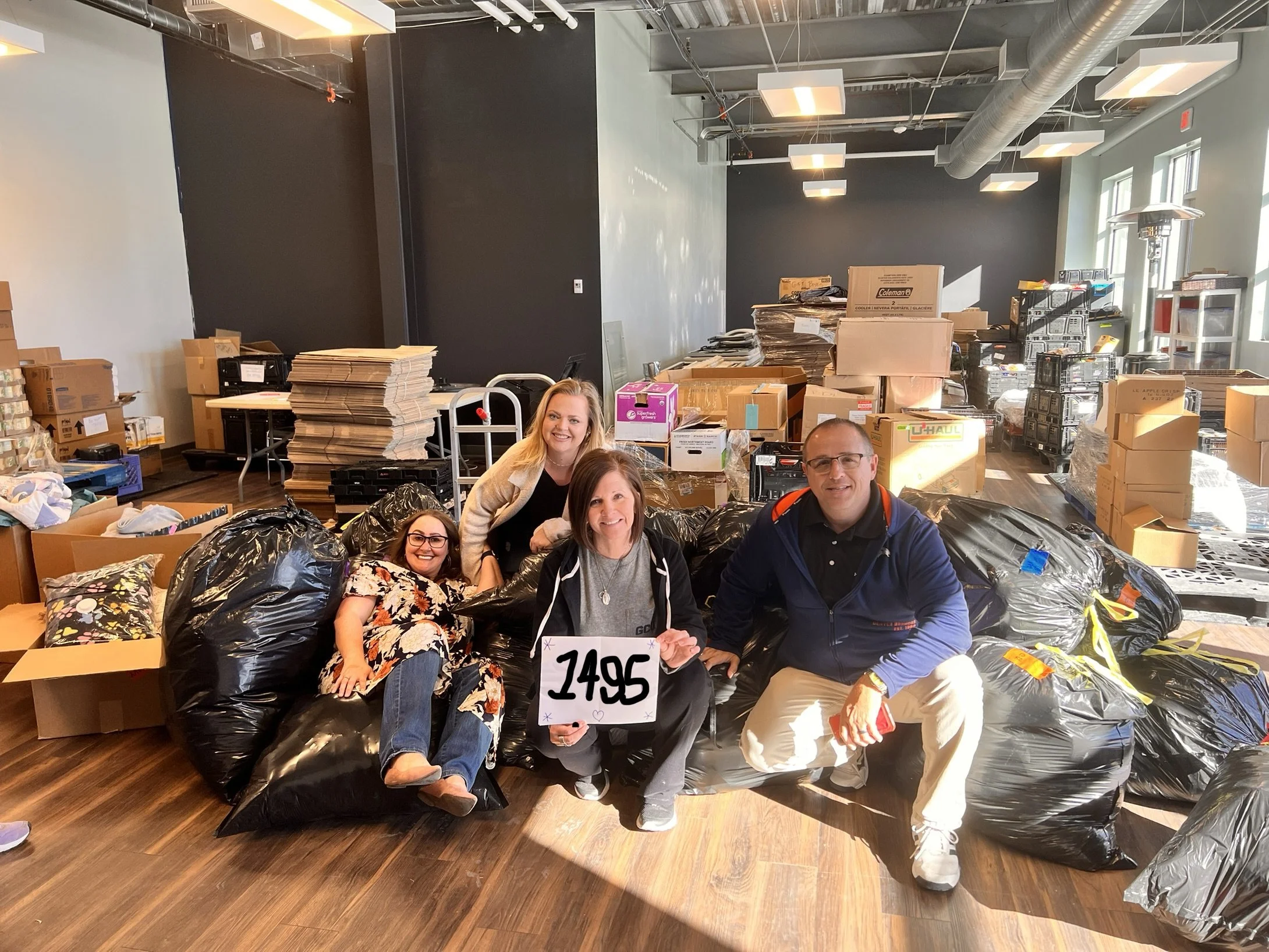 Four people smiling and posing with black trash bags and a sign with the number 1495 in a room filled with boxes, packages, and clutter, likely during a donation or packing event.