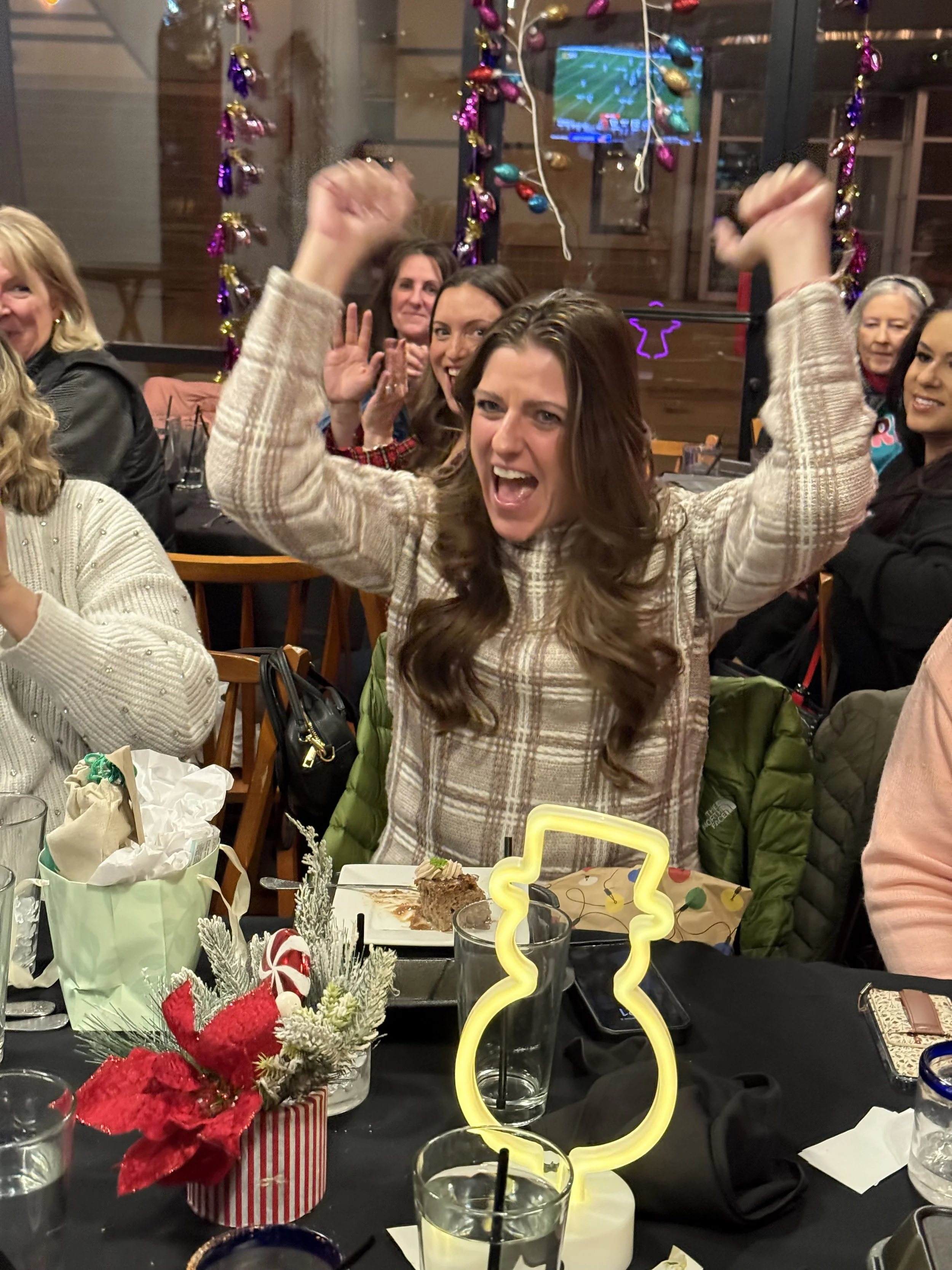 Woman celebrating with arms raised, surrounded by friends at a festive dinner party with holiday decorations and a cake.