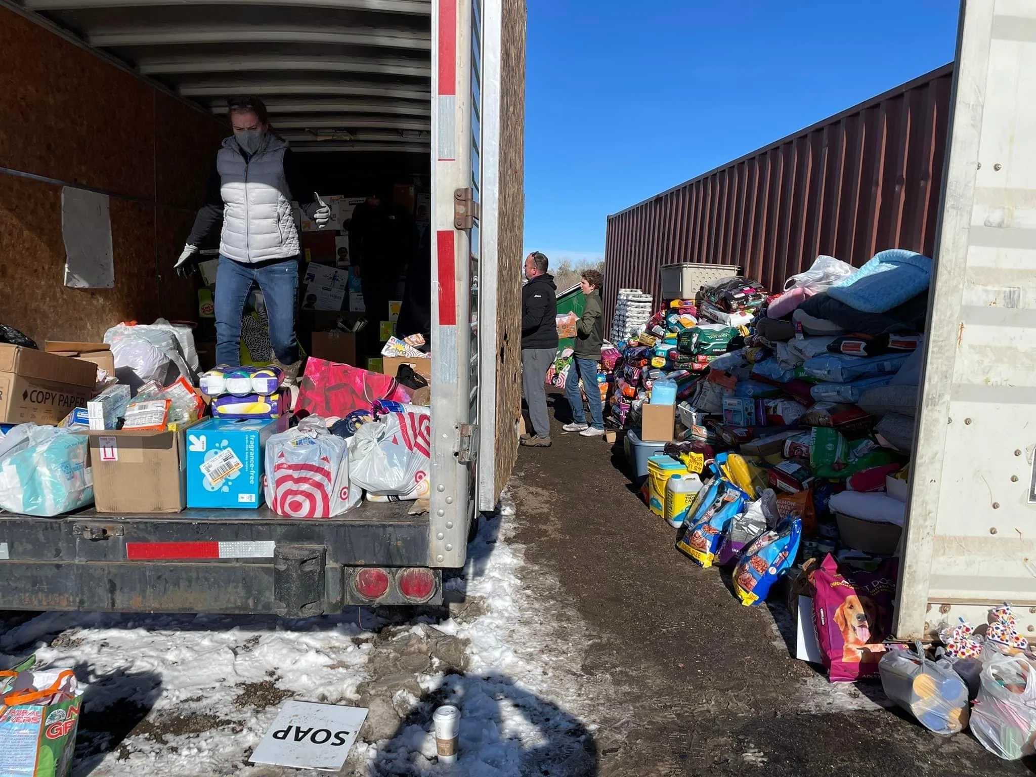 People unloading and sorting donated supplies at a community collection site, with items such as blankets, pet food, and household goods.
