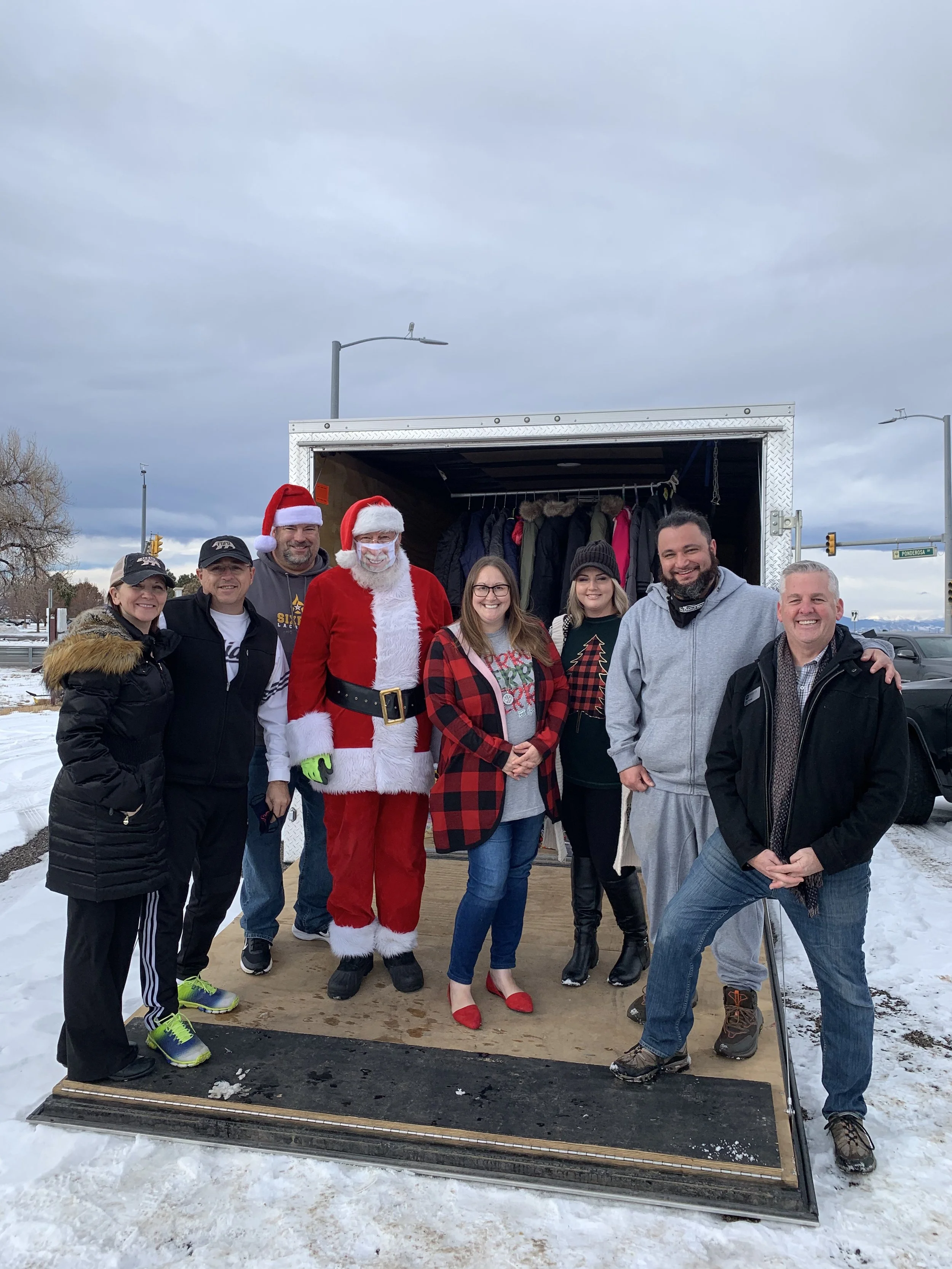 Group of eight people standing in front of a truck with coat rack inside, two dressed as Santa Claus, outdoors on snowy day.
