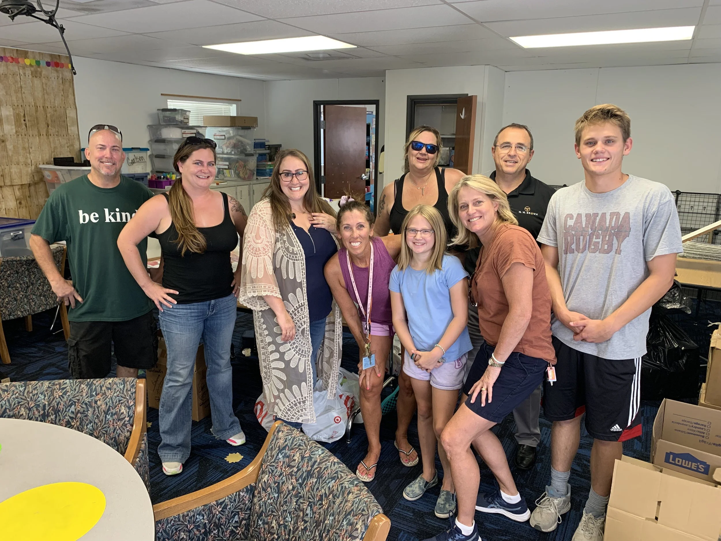 Group of ten people gathered in a room, smiling for the photo. The room has boxes, shelves, and a table with chairs with patterned upholstery. The group includes men, women, and children, dressed casually.