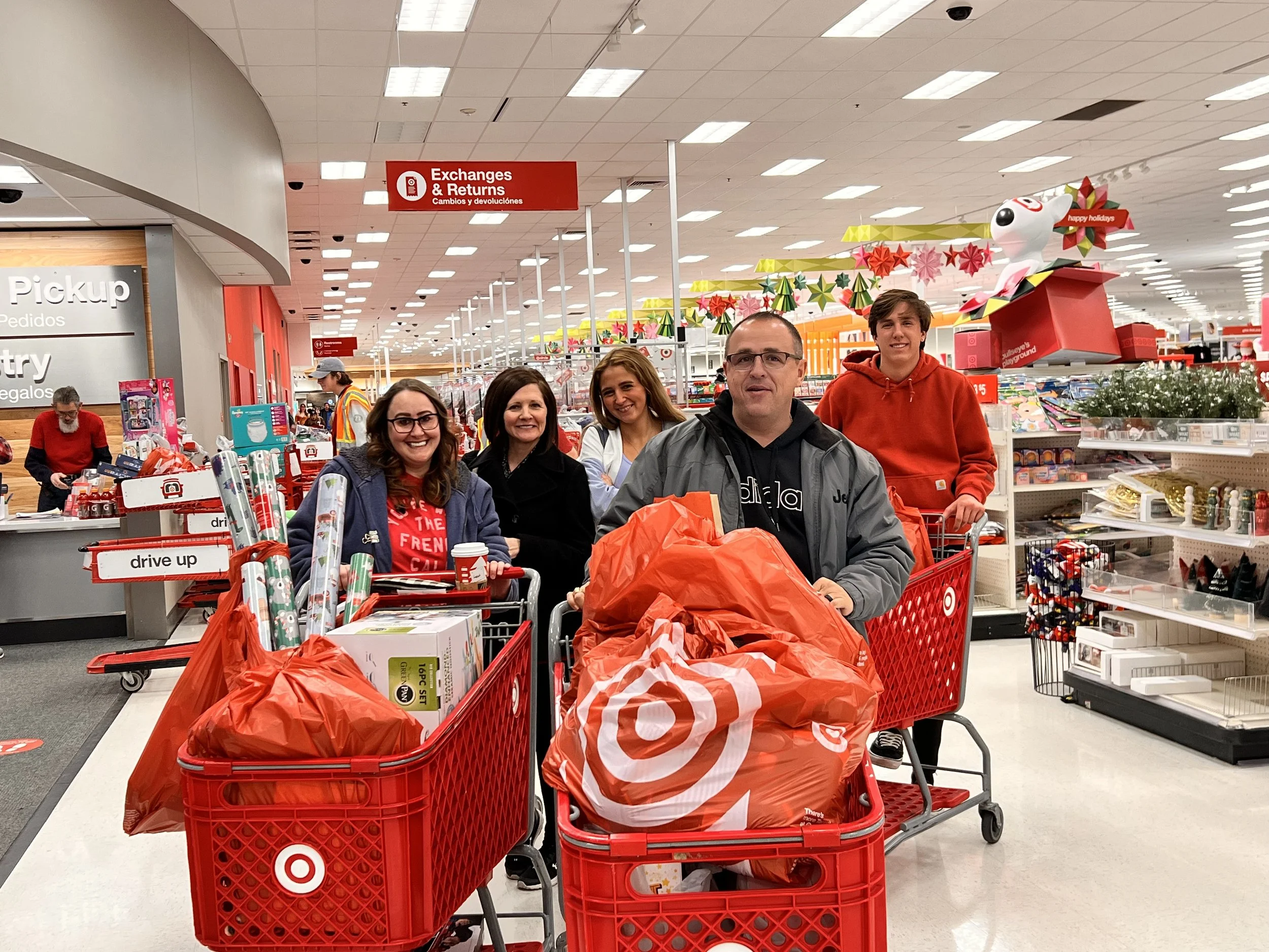 A group of five people standing with shopping carts filled with Target purchases in a store aisle, decorated for the holidays with red and green decorations, a Target logo on the carts, and Christmas-themed displays.