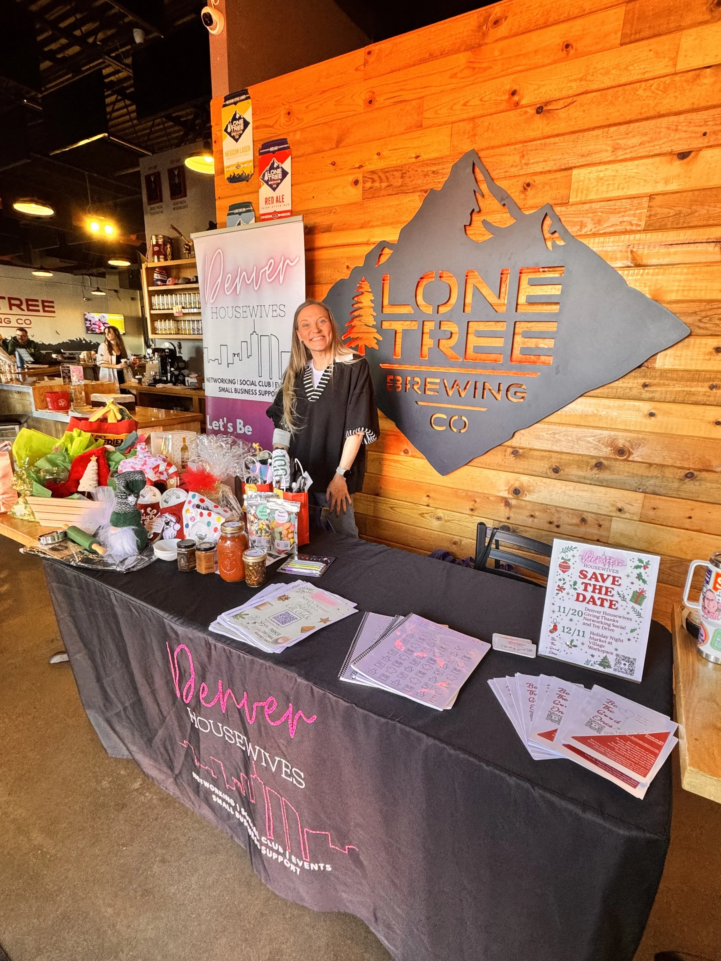 Woman standing behind a table at a booth for Denver Housewives Networking event, with a large sign for Lone Tree Brewing Co. on a wooden wall background, and promotional materials and holiday decorations on the table.