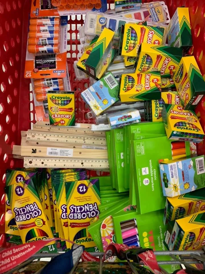 A red shopping cart filled with school supplies, including boxes of Crayola crayons, glue sticks, rulers, colored markers, and other stationery items.