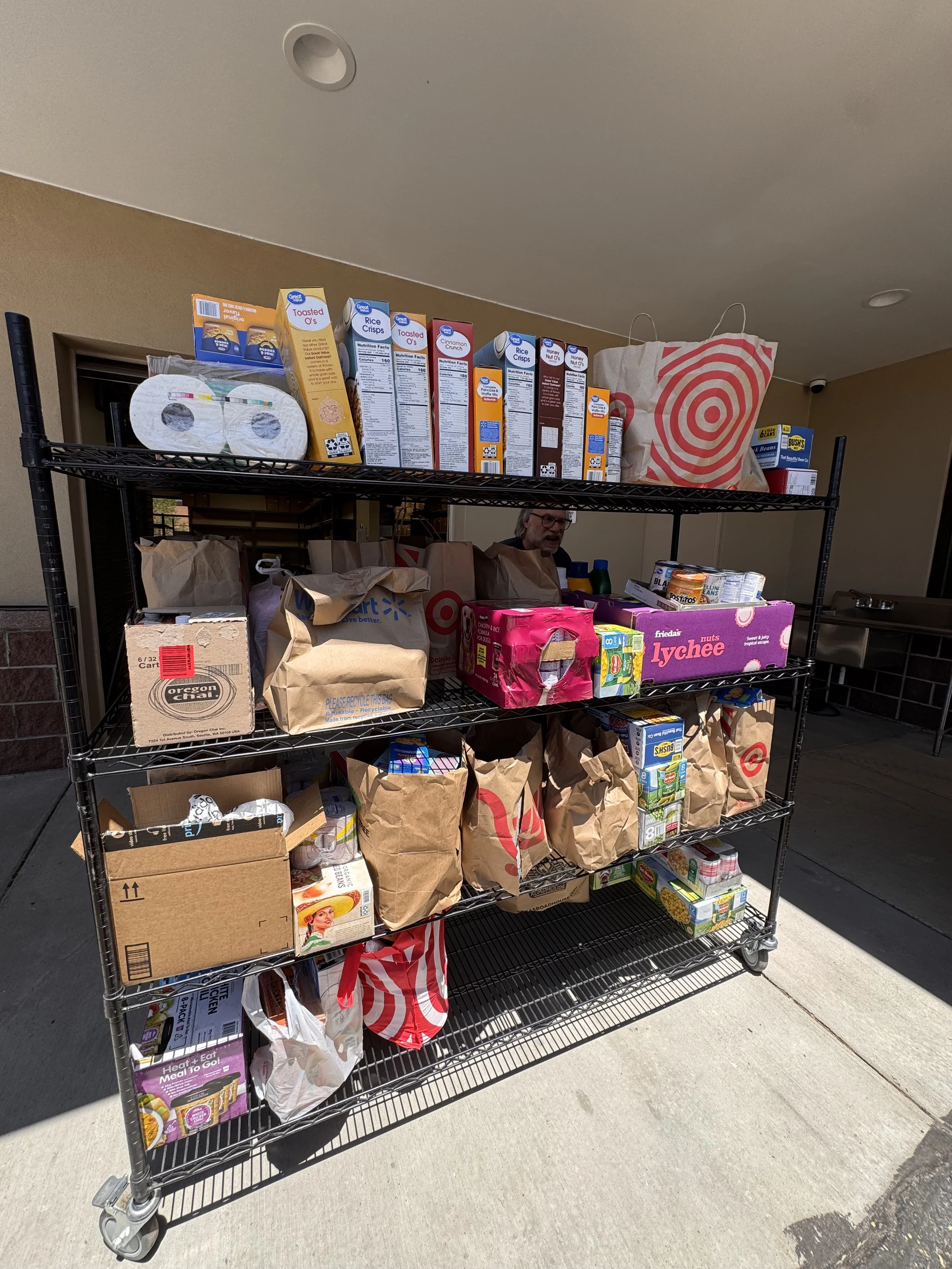 A metal cart filled with grocery bags and boxes outside a building, with some people visible behind it.