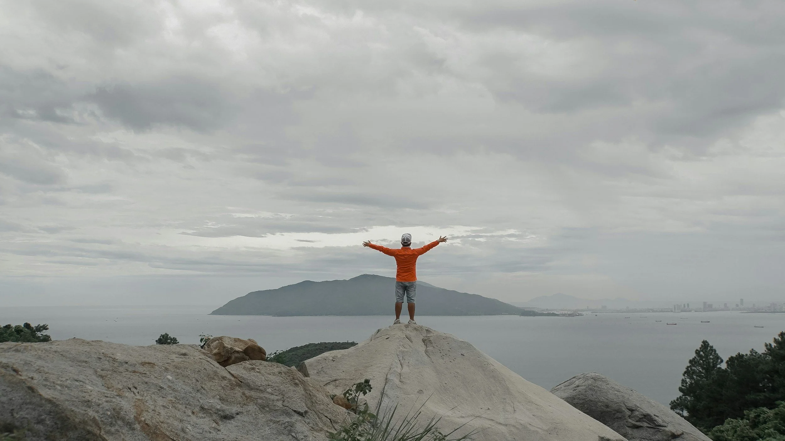 Person standing on a large rock with arms outstretched, overlooking a body of water and distant hills or mountains under cloudy sky.