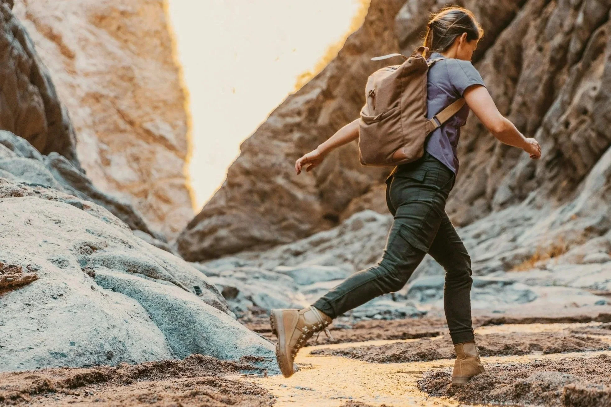 Woman jumping over a stream onto a sandy bank during a hike through a rocky canyon with backpack.