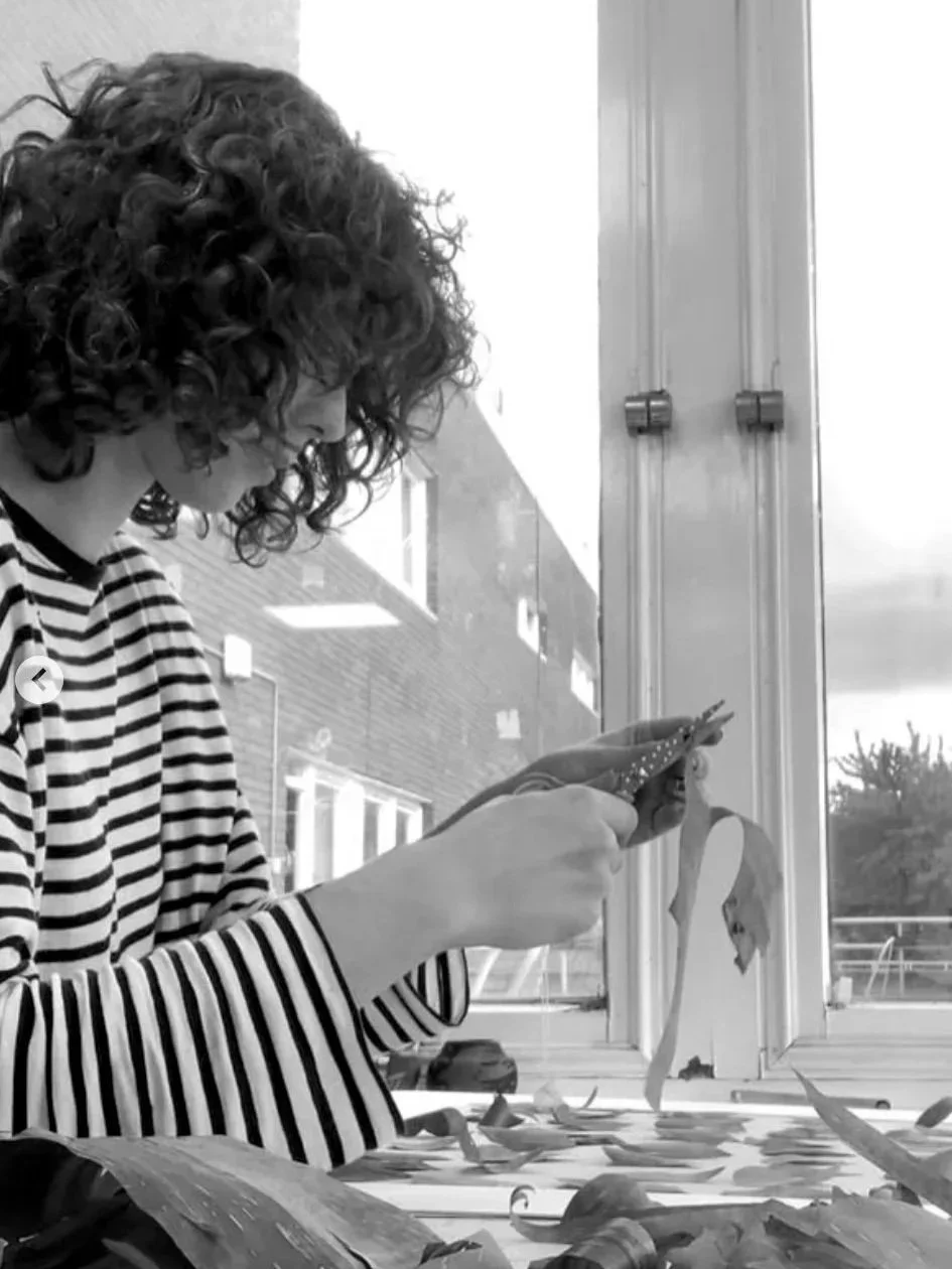 A person with curly hair and wearing a striped shirt is sitting by a window, making a collage on the table.