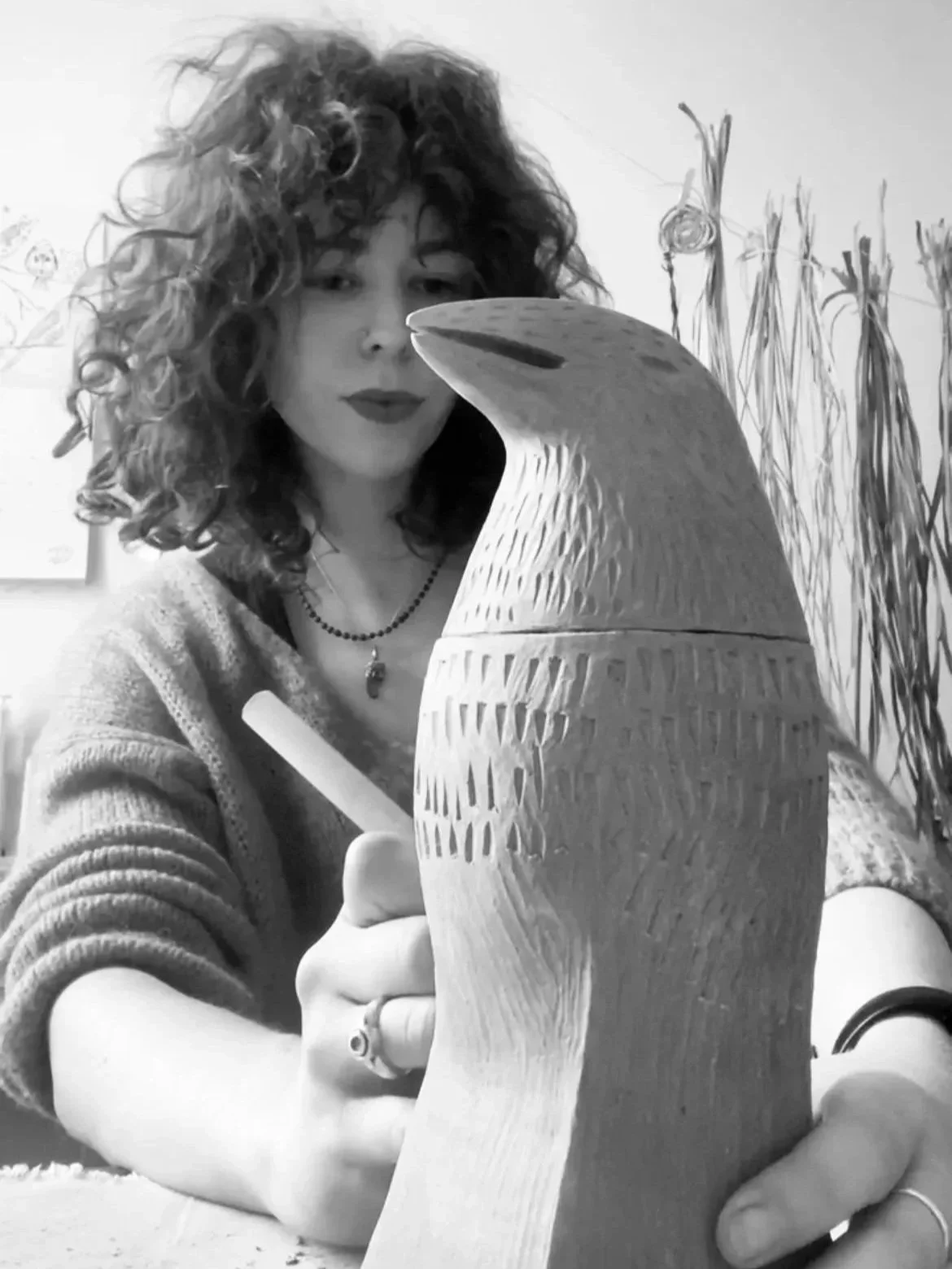 A woman with curly hair and a necklace is drawing or carving a large wooden sculpture that resembles a bird's head. She is using a tool to work on the sculpture and appears focused.