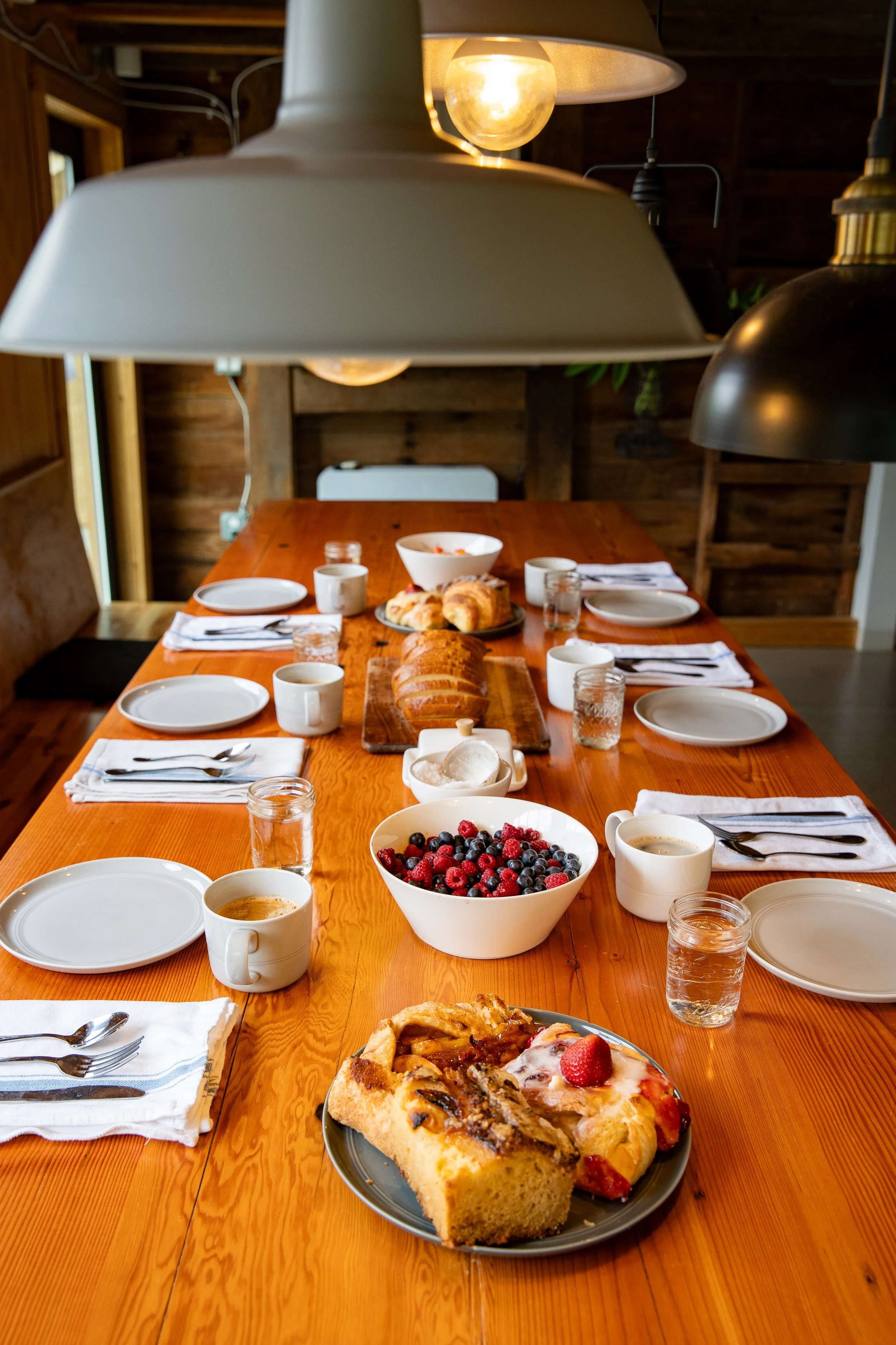 A wooden dining table set with plates, cups of coffee, glasses of water, and an assortment of breakfast foods including a bowl of mixed berries, slices of bread, pastries, and slices of cake, under hanging lamps in a cozy wooden room.