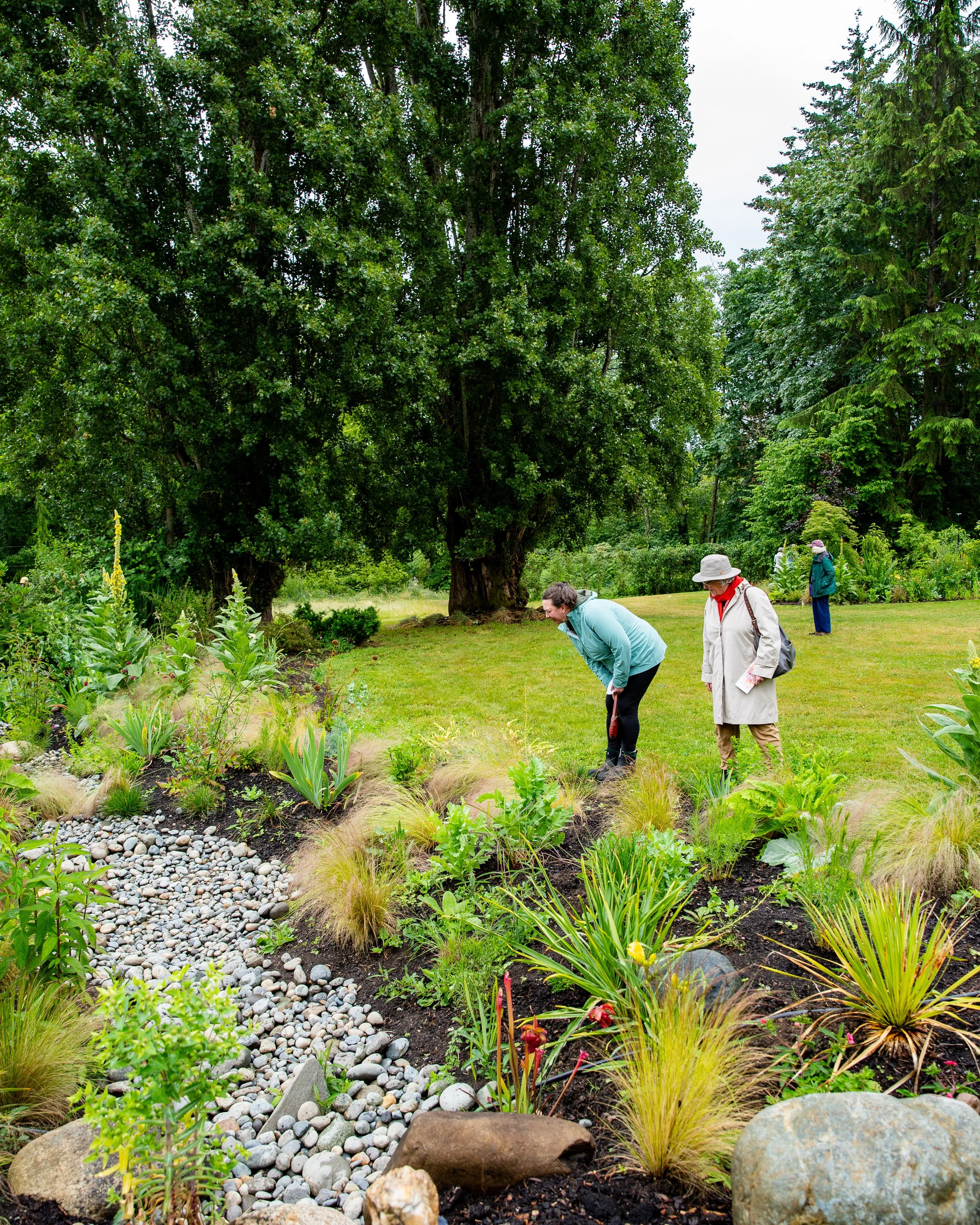 Three people gardening in a lush, green park with mature trees and well-maintained flower beds.
