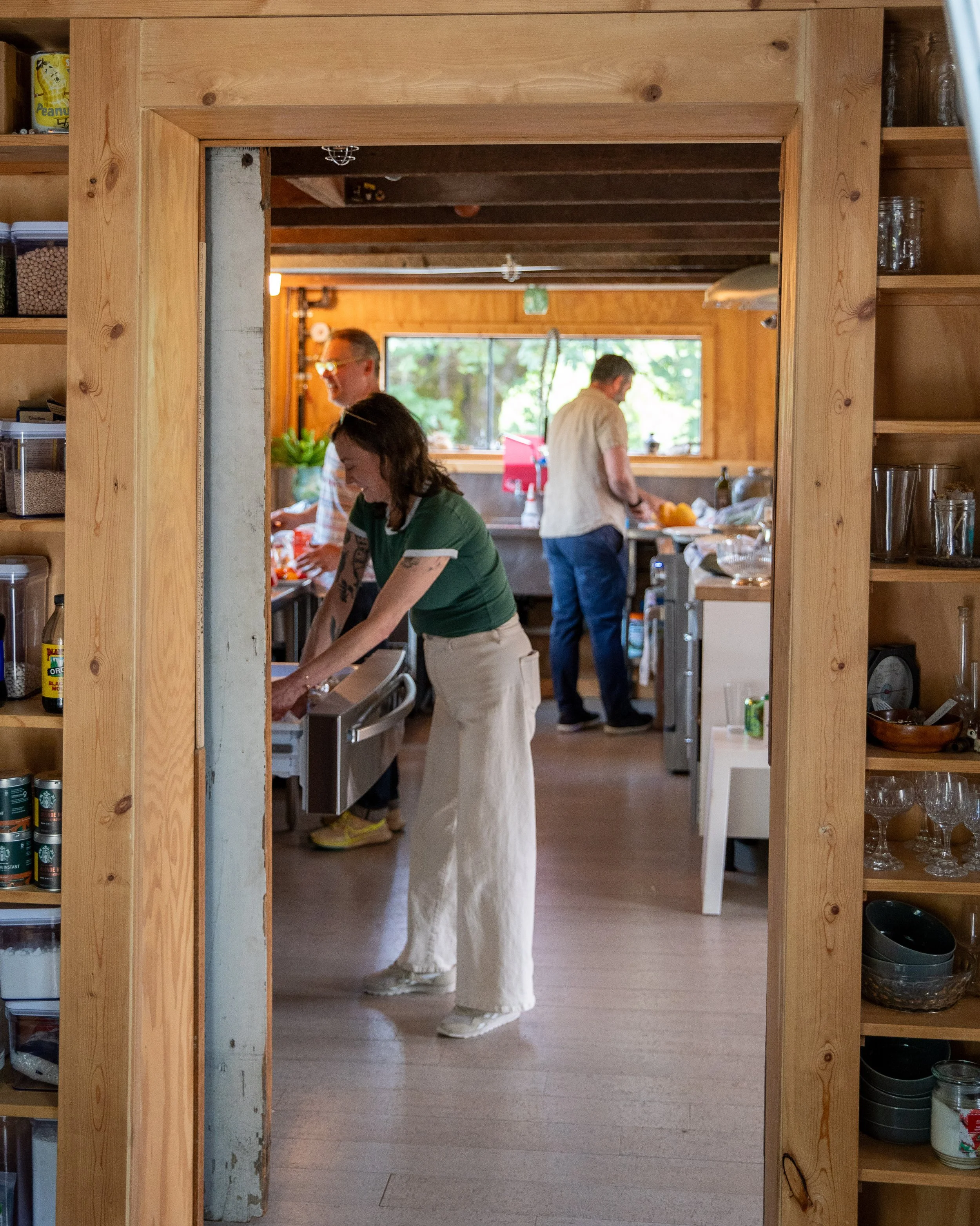 Three people preparing food in a rustic kitchen with wood-paneled walls and large windows, seen through a doorway with wooden framing.