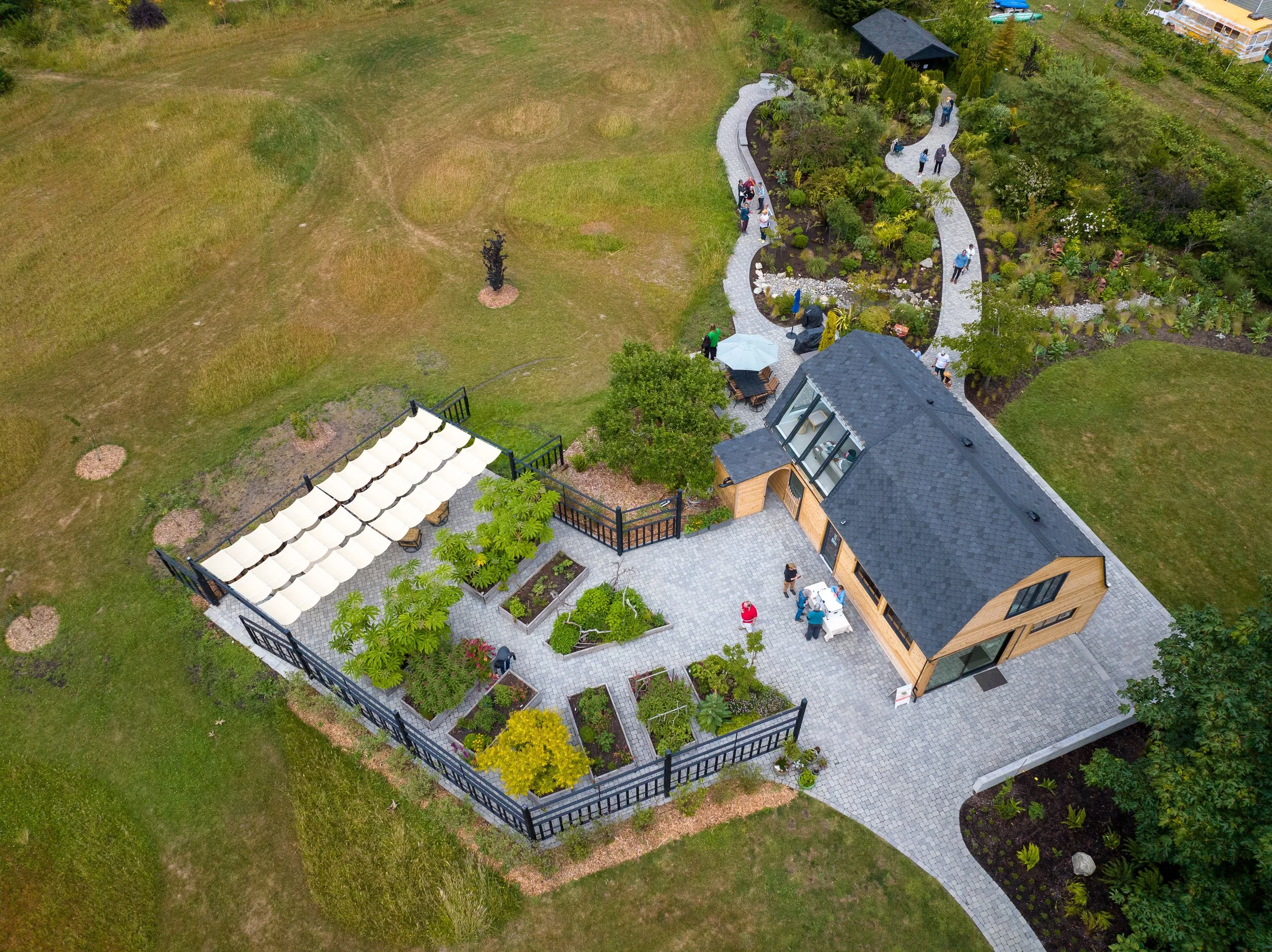 Aerial view of a modern house with a garden, patio, and a winding pathway filled with people, surrounded by grass and trees.