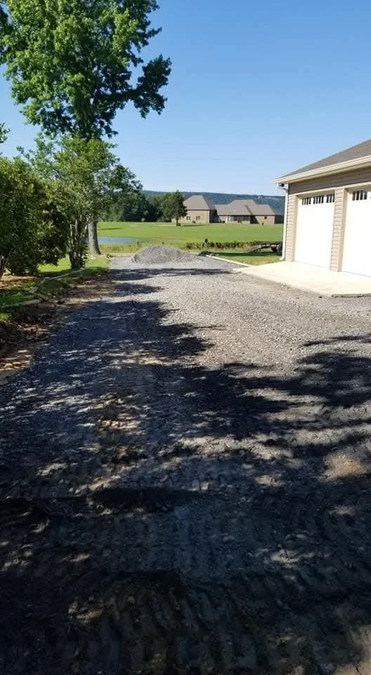 A gravel driveway next to a garage with a closed white door, side yard with trees, green lawn, and houses in the background under a blue sky.