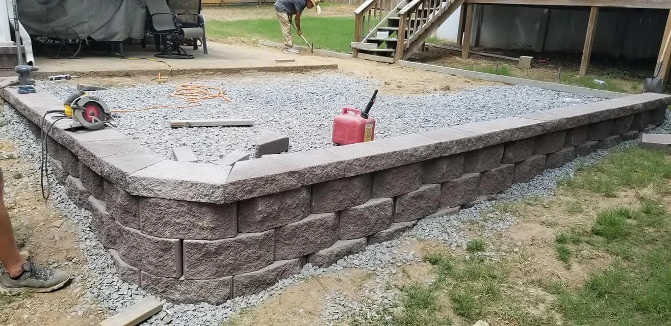 Construction of a stone retaining wall with large, rectangular granite blocks in a backyard. A worker is seen in the background working on the project, with tools and materials like a saw and a red fuel container nearby.