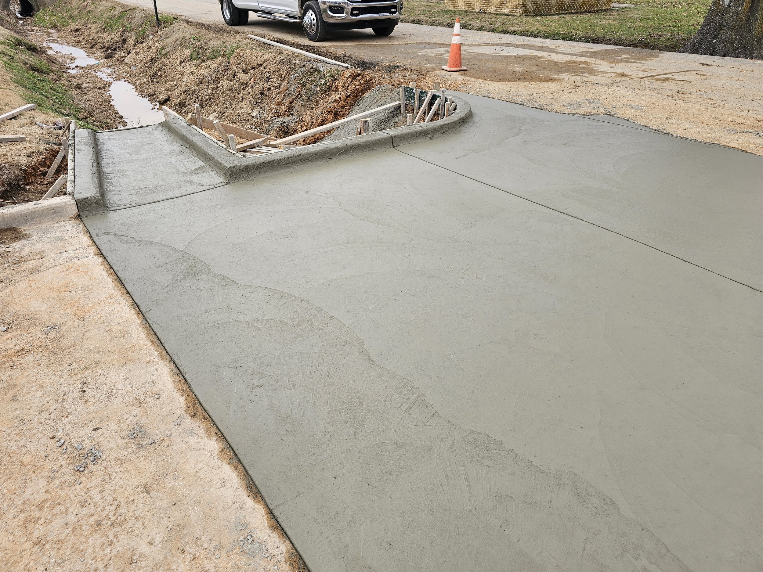 Construction site with new concrete sidewalk, orange traffic cone, and a silver pickup truck on the road