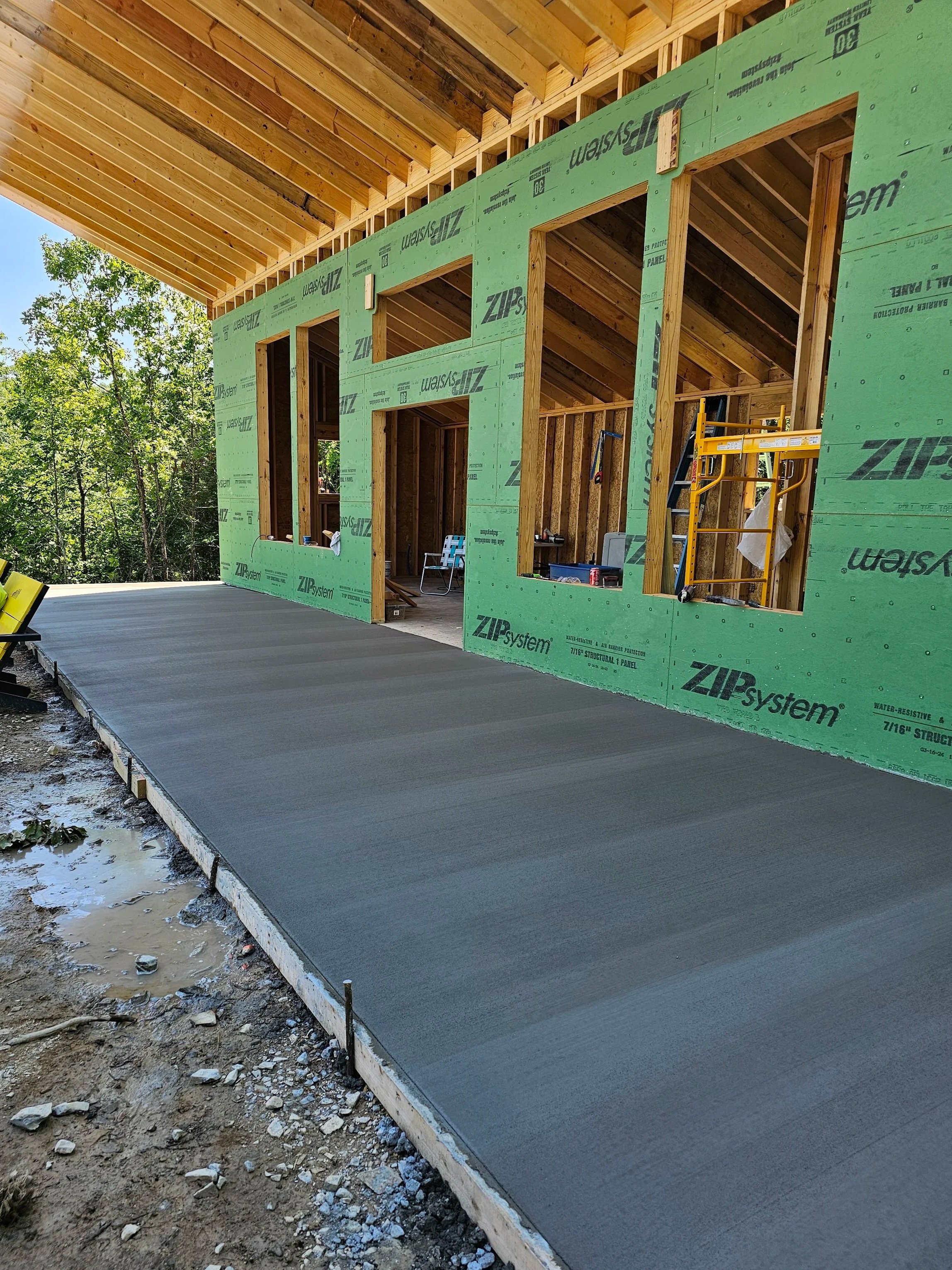 Construction site of a house with a freshly poured concrete slab, unfinished wooden framing, partially installed sheathing, and tools and a scaffold inside.