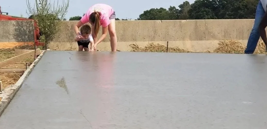 Two children and an adult working on pouring and smoothing concrete on a flat surface outdoors.