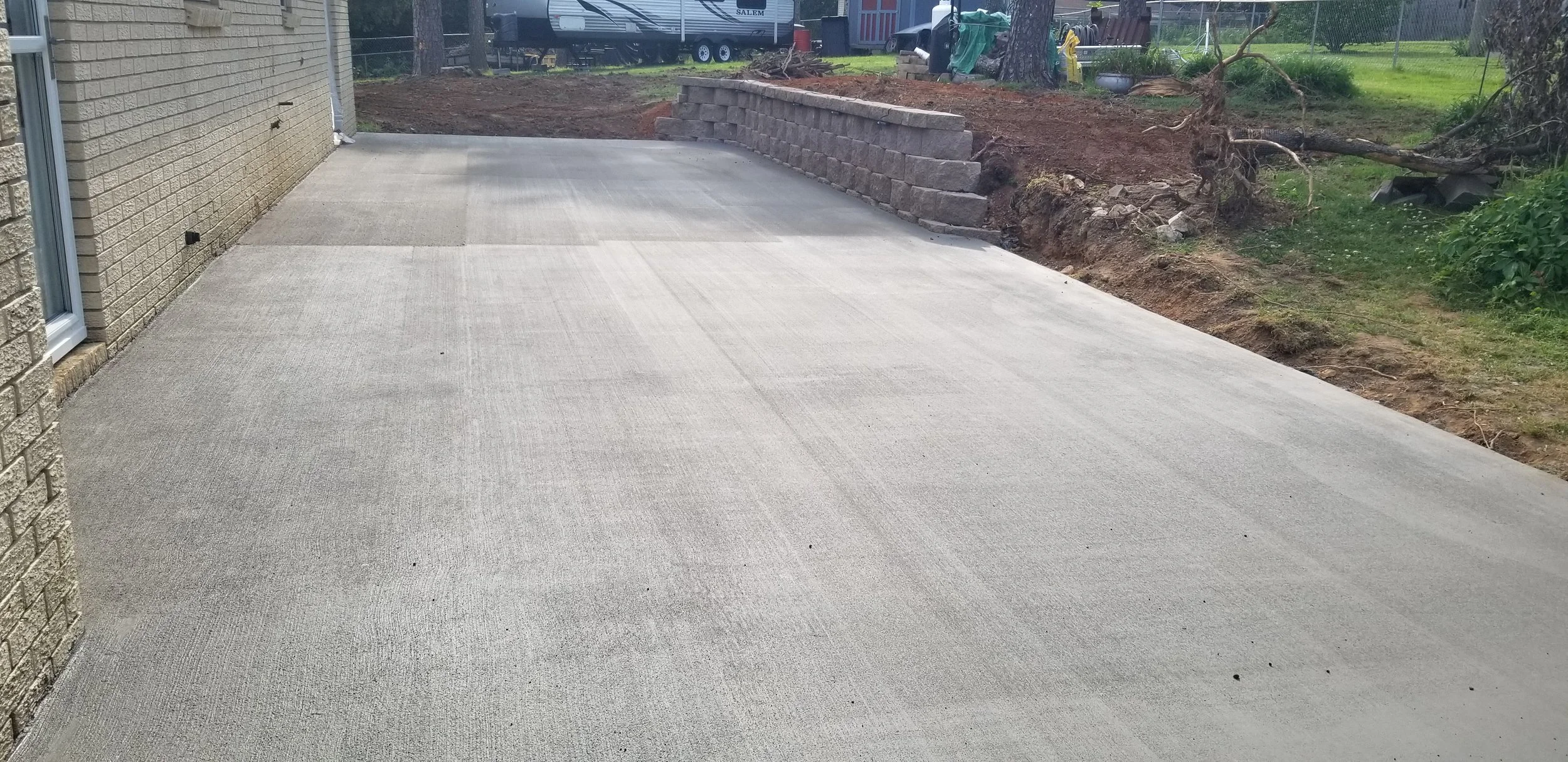 Newly poured concrete patio with a retaining wall of bricks on the edge, green yard with trees, and construction materials in the background.