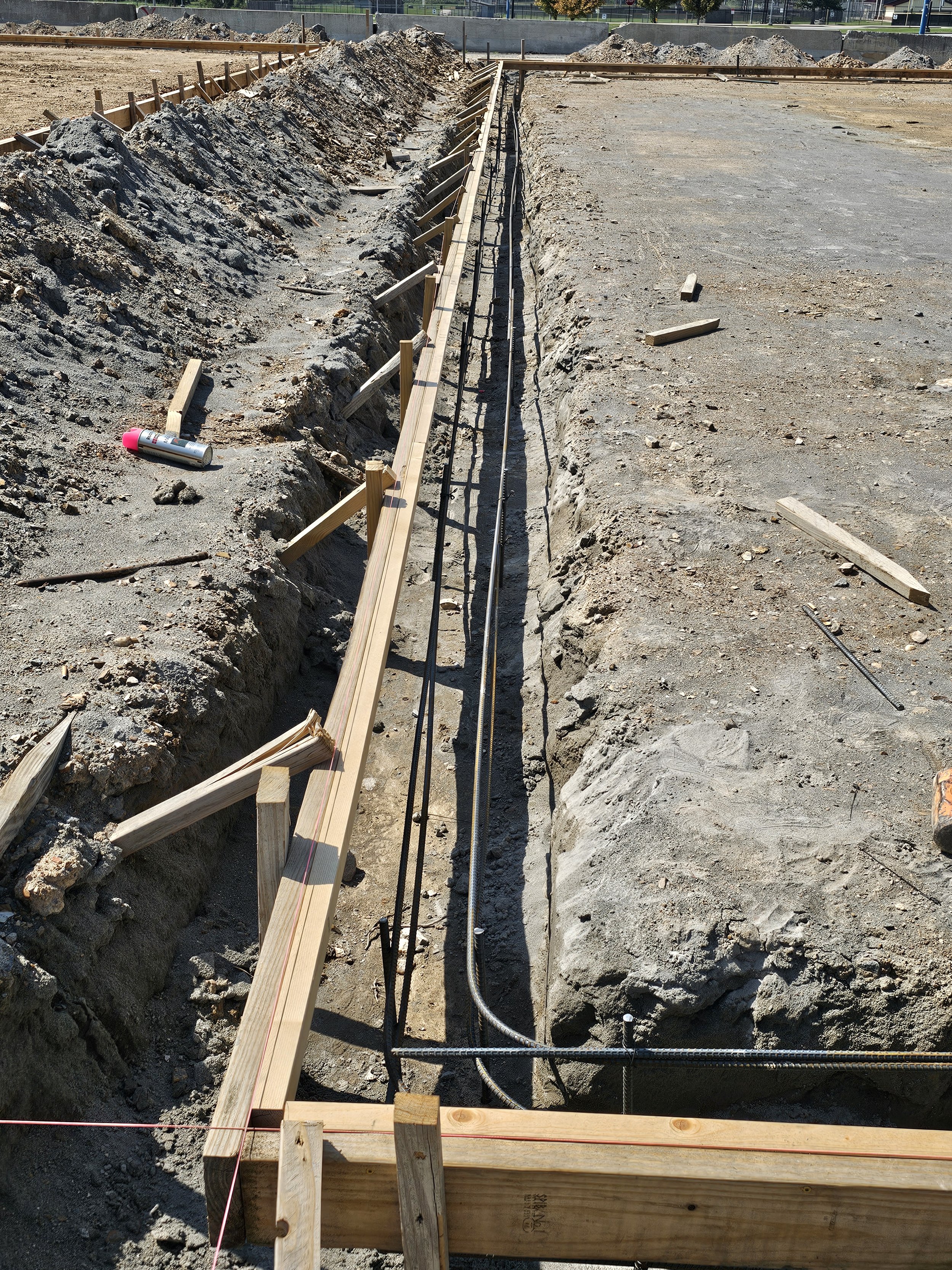 Construction site with trench dug in ground, wooden and metal supports, and construction tape. The trench appears to be for installing underground utilities or piping.