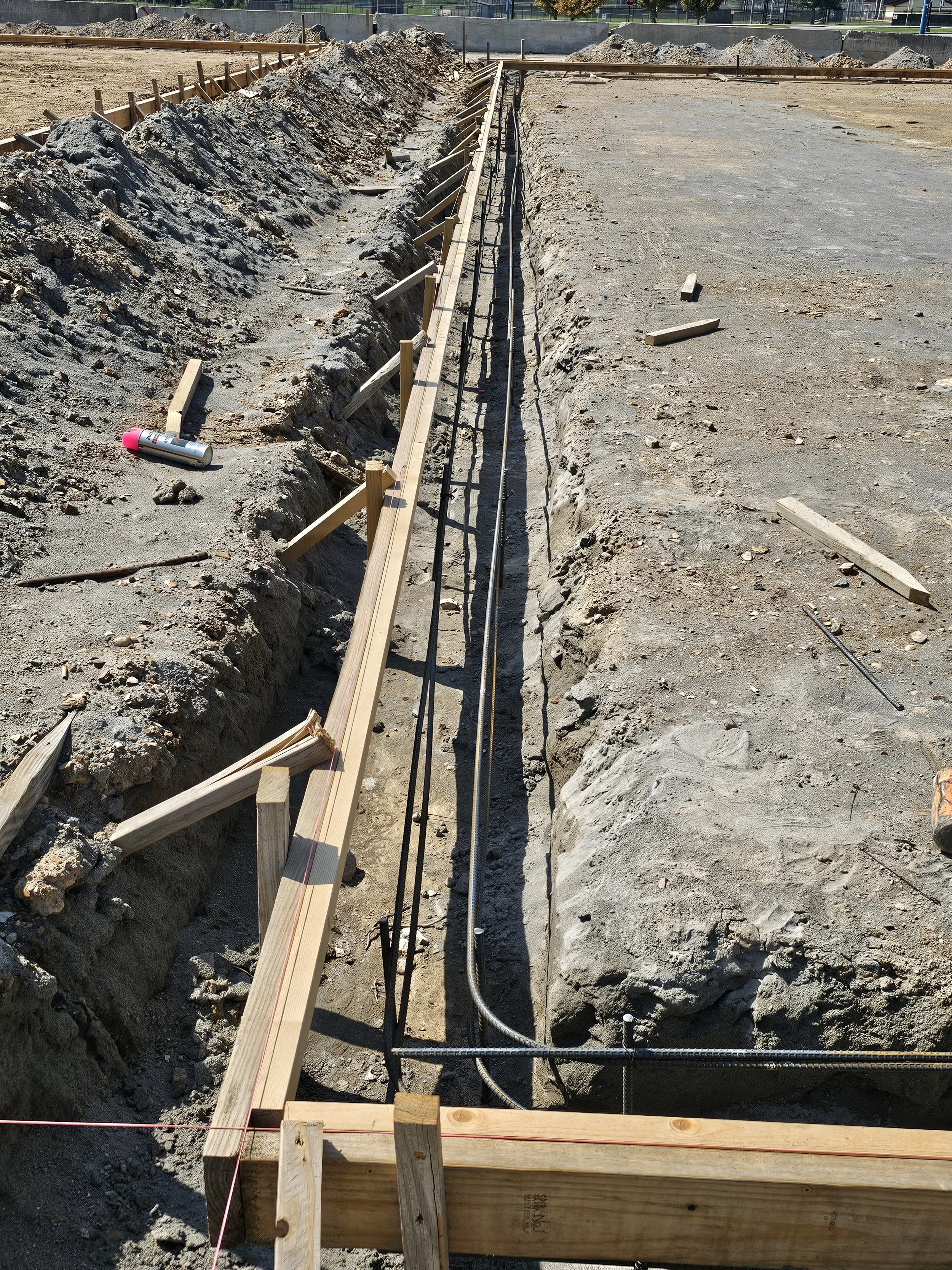 Construction site with trench dug in ground, wooden and metal supports, and construction tape. The trench appears to be for installing underground utilities or piping.