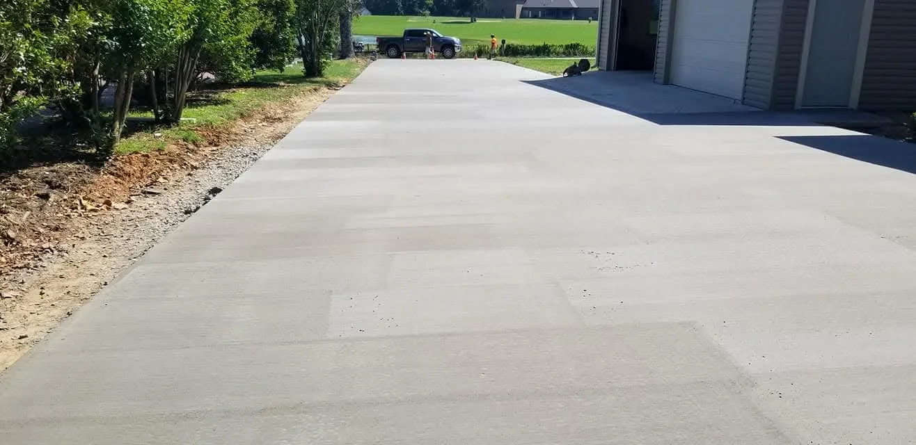 Freshly poured concrete driveway extending from a garage to the street, with a pickup truck and a few cars parked in the distance.