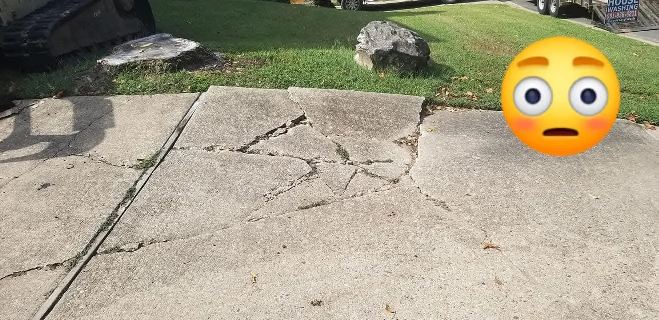 Cracked sidewalk with visible damage and two large rocks in the background, grass and a vehicle partially visible.
