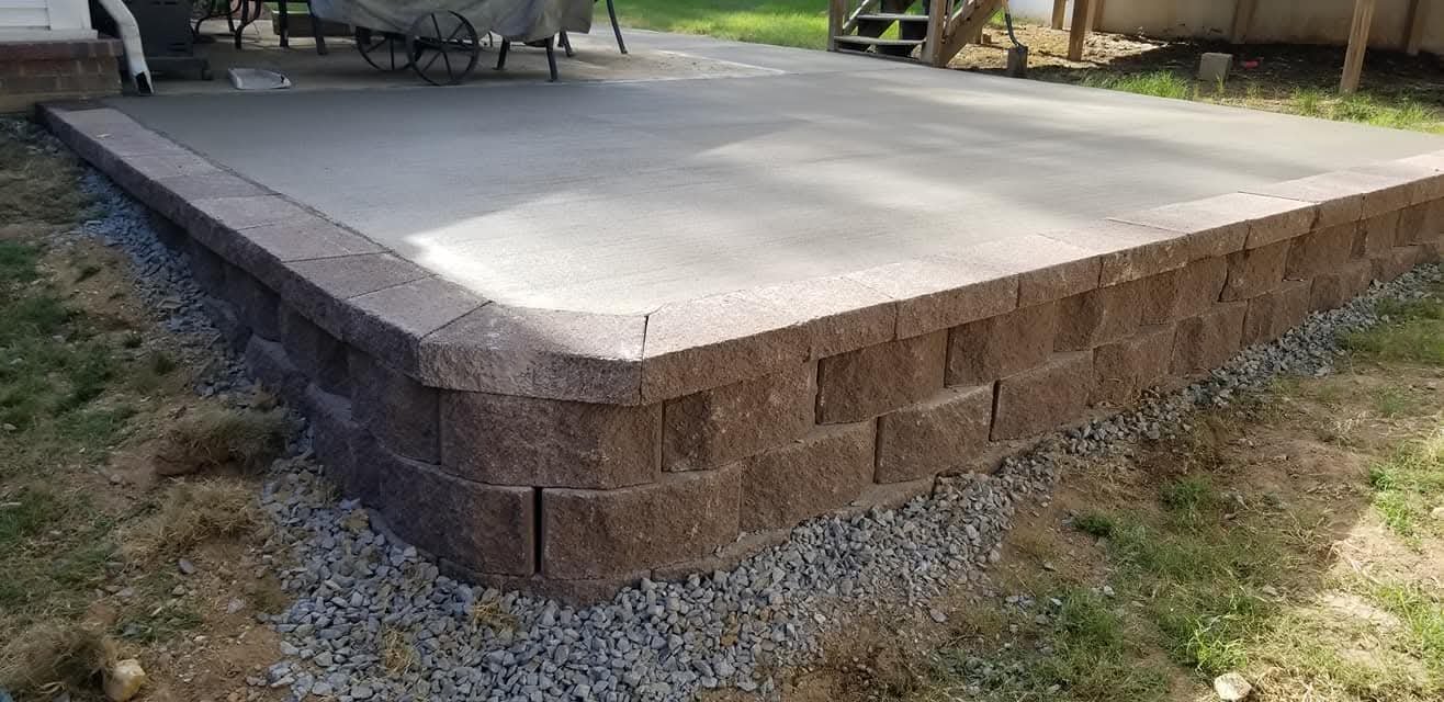 Close-up view of a newly constructed stone patio with a slight kick at the corner, elevated on a stacked stone wall with gravel underneath, outdoor setting with grass and wooden stairs in the background.