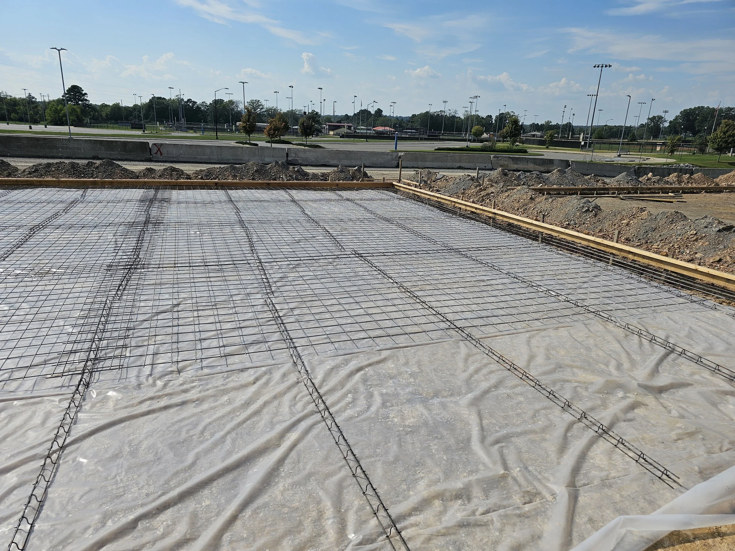 Construction site with rebar and concrete foundation, surrounded by dirt and a concrete barrier, with a tennis court and trees in the background under a partly cloudy sky.