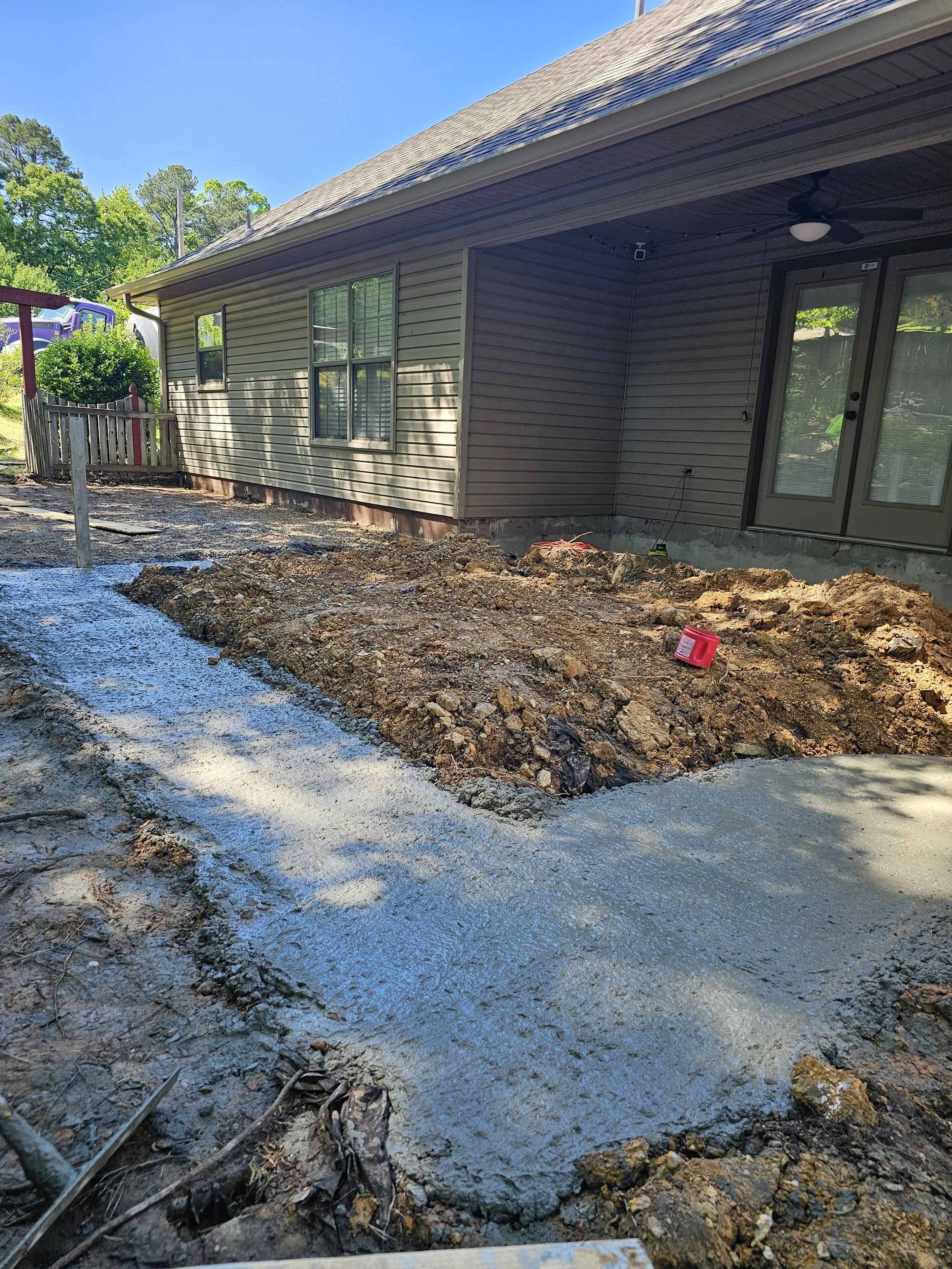 Photo of an exterior house with beige siding, a covered porch, and a sliding glass door. The ground in front is under construction, with dirt, gravel, and mud, possibly for landscaping or paving. There is an overhead ceiling fan on the porch and some