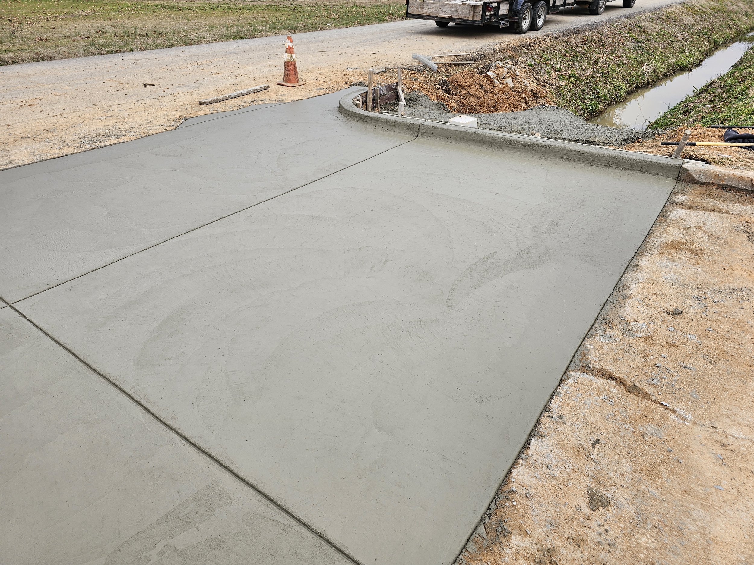 Finished gray concrete sidewalk next to a small ditch with water and dirt, construction tools and safety cones nearby, outdoor setting during daytime.