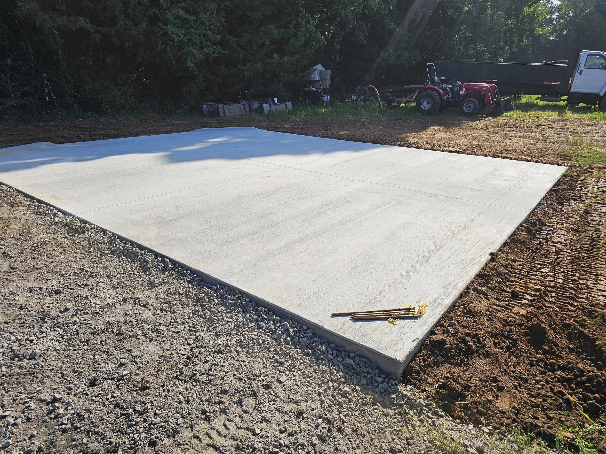 Freshly poured concrete slab on a construction site, with tools placed on its edge, surrounded by dirt and gravel, with a trailer and machinery in the background.