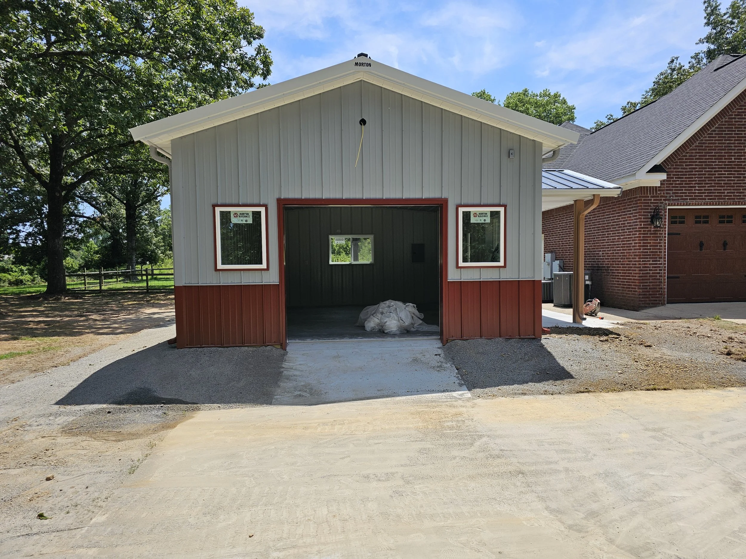 A partially constructed garage or shed with gray and red siding, two windows, and an open garage door, situated next to a brick house; trees and a blue sky in the background.