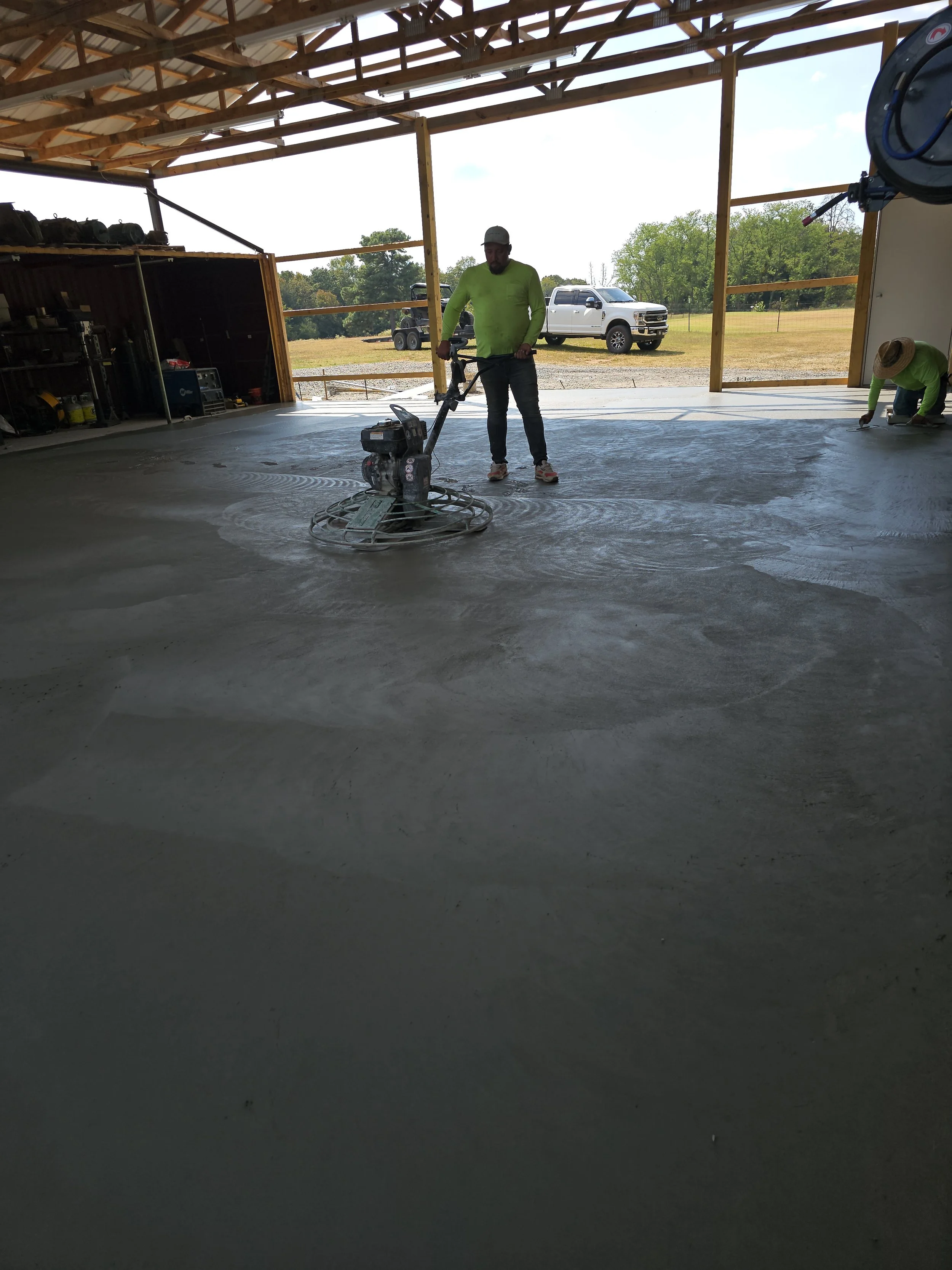 A person in a green long sleeve shirt and a hat is using a power trowel to smooth a concrete floor inside an unfinished building with open walls and a view of outdoor grass and trees.