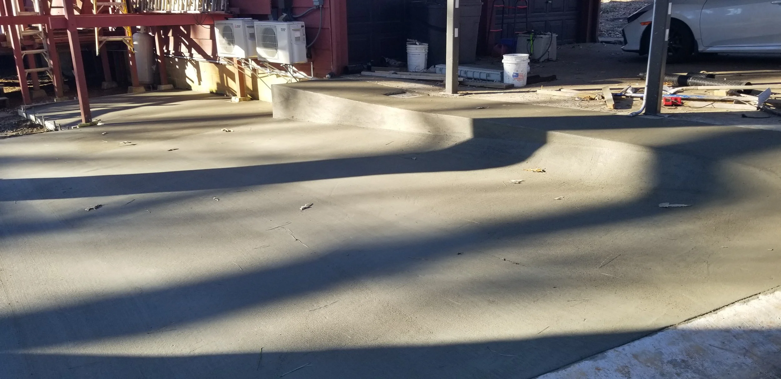 Construction site with fresh concrete sidewalk, stairs, construction equipment, and nearby parked car under sunlight.