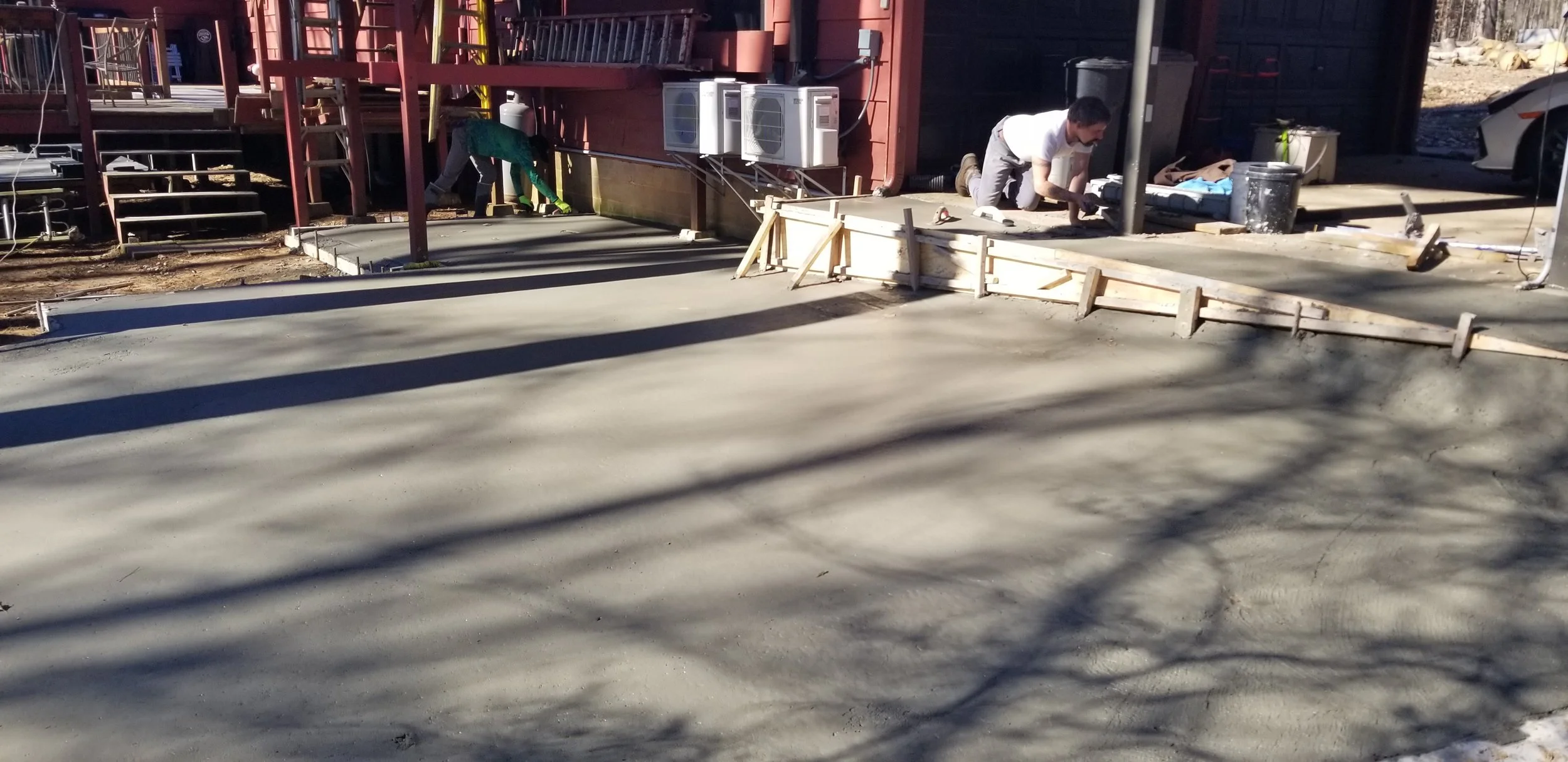 Construction workers pouring and smoothing concrete for a sidewalk in front of a building with red siding, stairs, and air conditioning units.