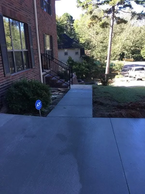 Concrete sidewalk leading to stairs with black railing near a brick building, with shrubs and a yard on the side, and parked cars in the background.