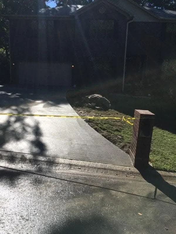View of a residential driveway and front yard with yellow caution tape across the yard, a brick mailbox, and a dark house in the background, with sunlight creating shadows.