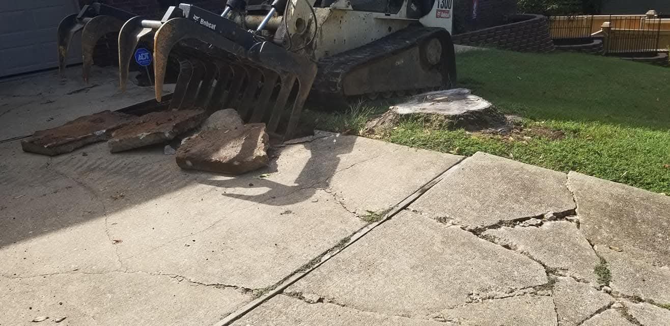 A small excavator is breaking a concrete sidewalk with large rocks and debris scattered around.