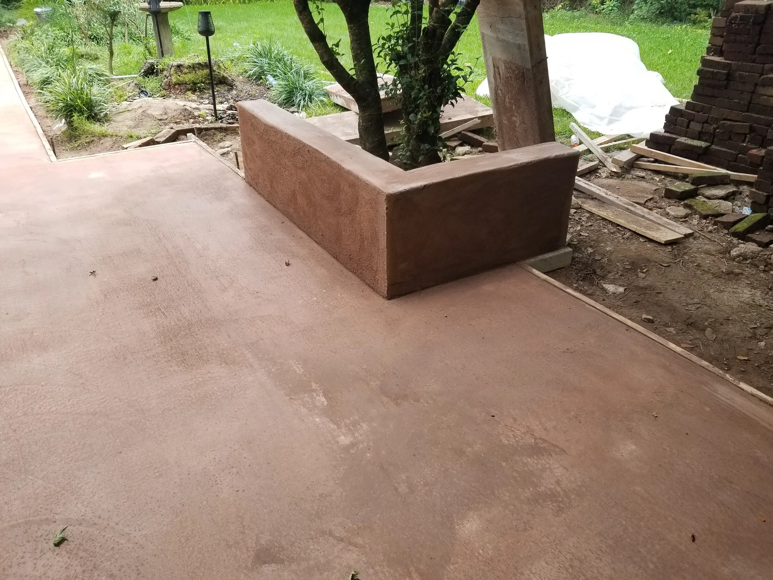 View of a backyard with a concrete patio, a brick-lined garden bed, trees, green grass, and tools scattered nearby, indicating ongoing construction or landscaping work.