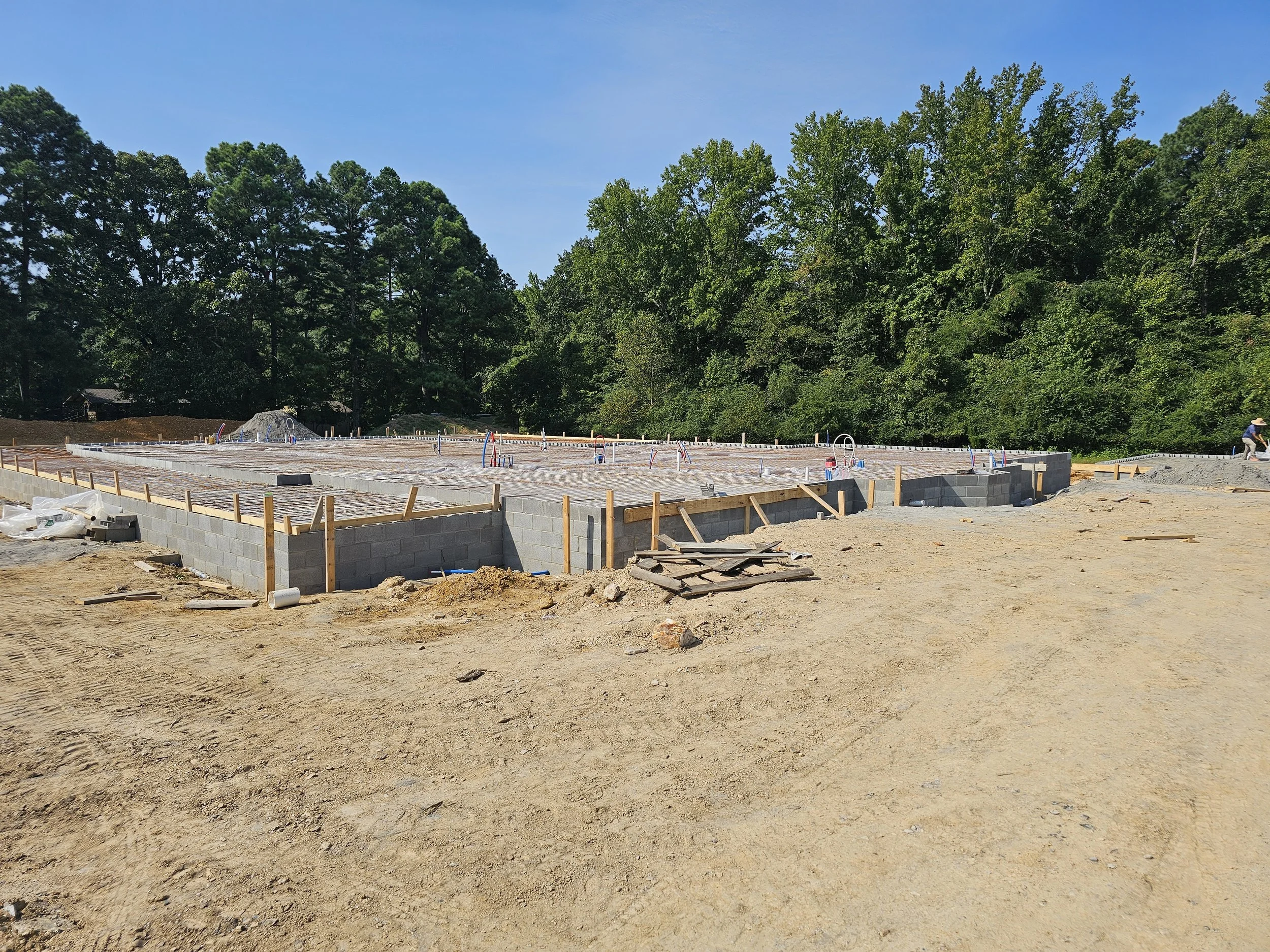 Construction site with a foundation in progress, surrounded by dirt and gravel, with trees and blue sky in the background.