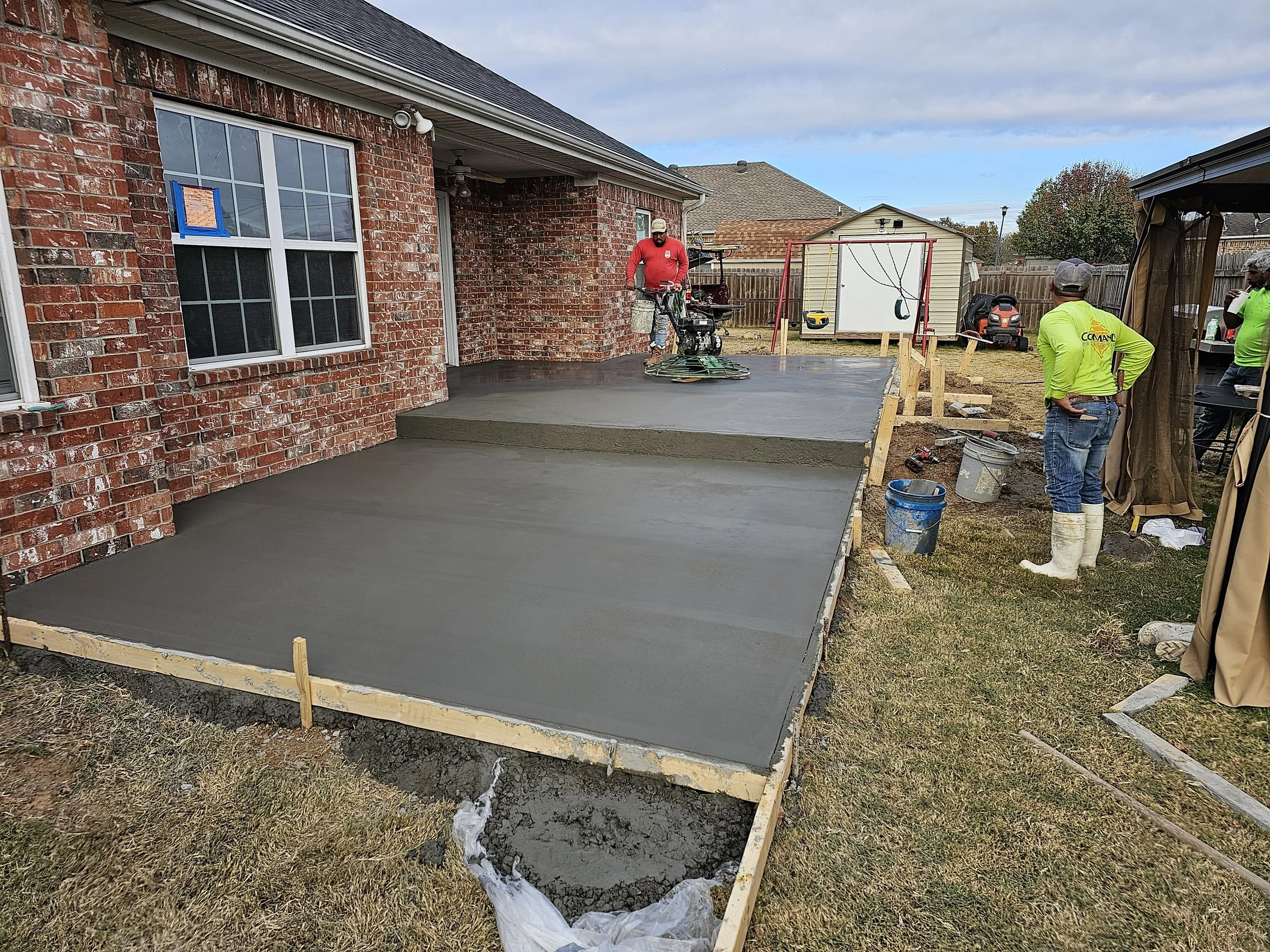 A backyard with a newly poured concrete patio. A person in a red shirt is operating a troweling machine on the concrete. Two people are observing, one in a bright yellow shirt and white boots, the other in a gray hat. Construction materials and tools