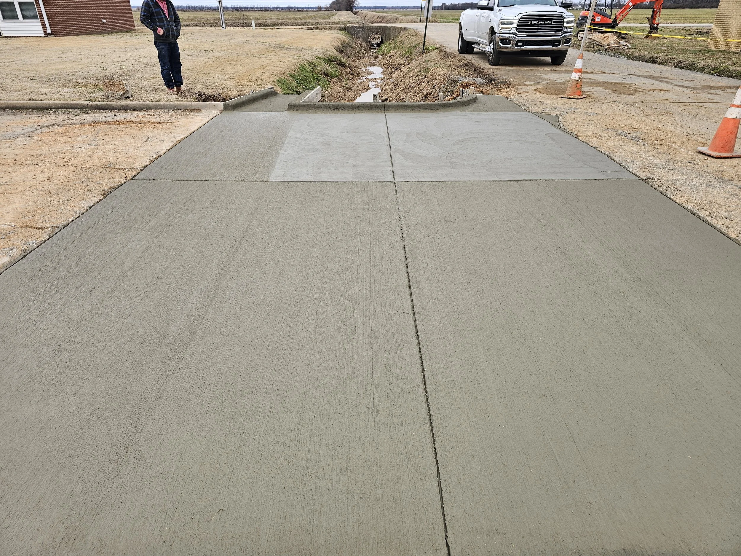 A newly poured concrete sidewalk in a rural area with construction cones and a man standing to the side. There are stopped vehicles and construction equipment in the background on overcast day.
