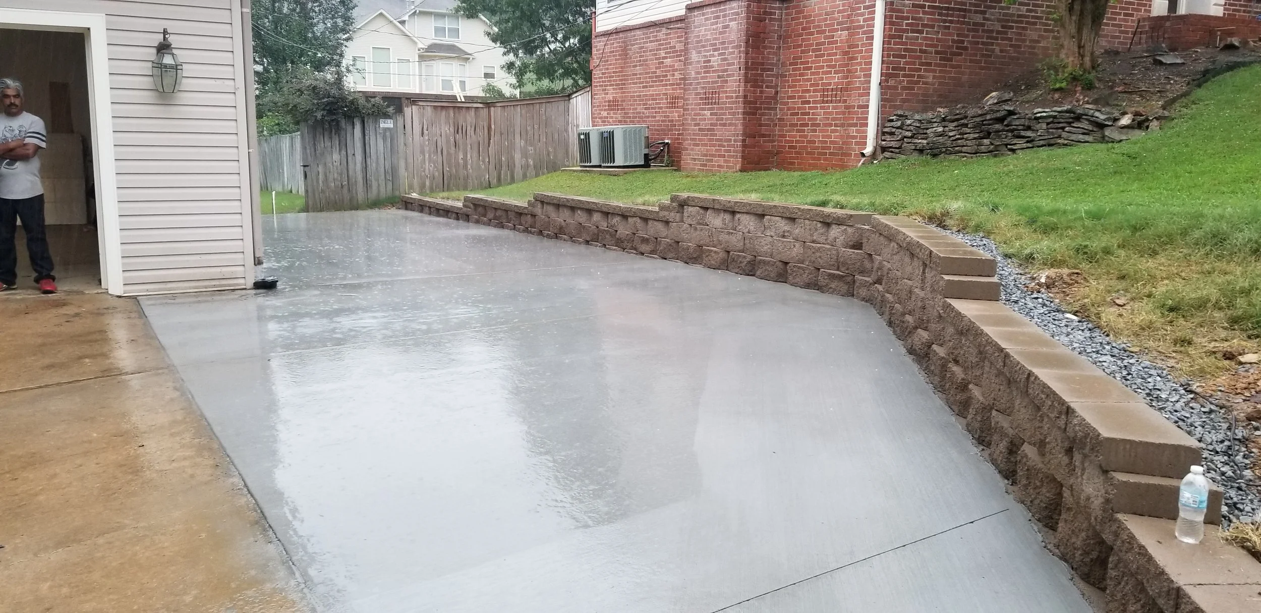 Wet concrete patio next to a house with a man standing in the doorway, a wooden fence, a brick wall, and a landscaped yard with grass and rocks.