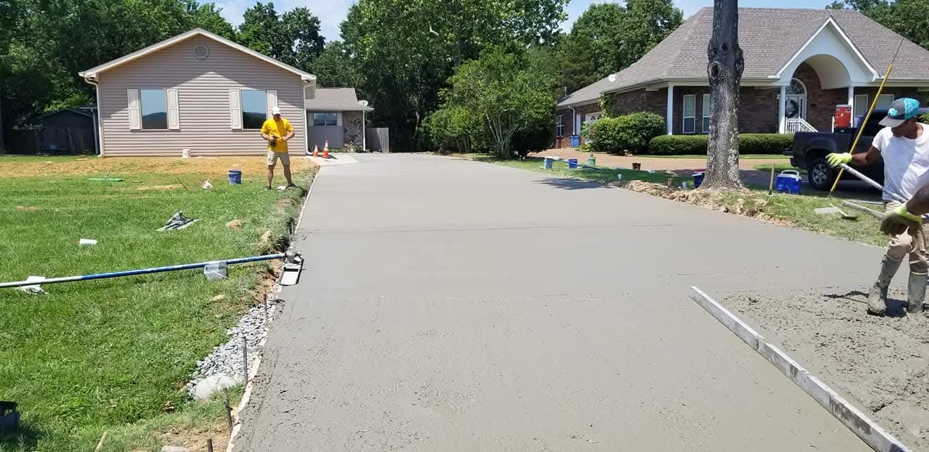 Workers are pouring and smoothing concrete on a residential driveway under a sunny sky.
