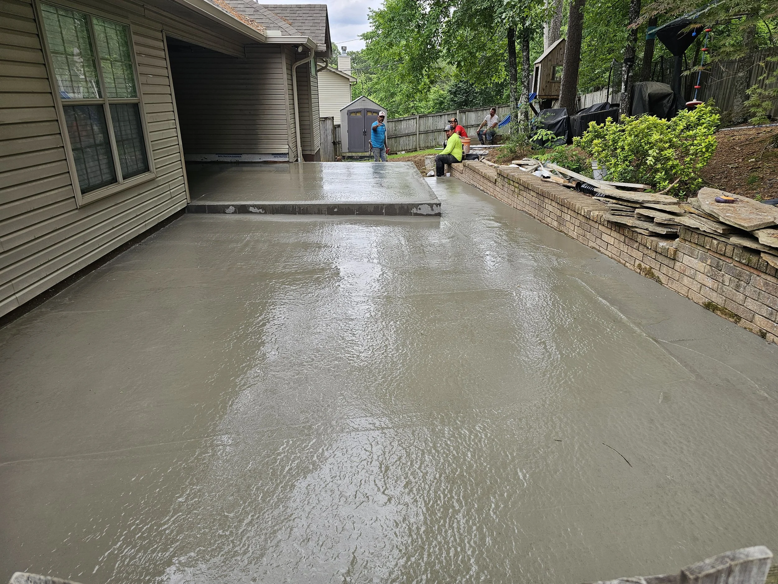 A group of five workers in bright safety clothing pouring and leveling concrete for a backyard patio. They are near a beige house with green trees and a wooden fence in the background.