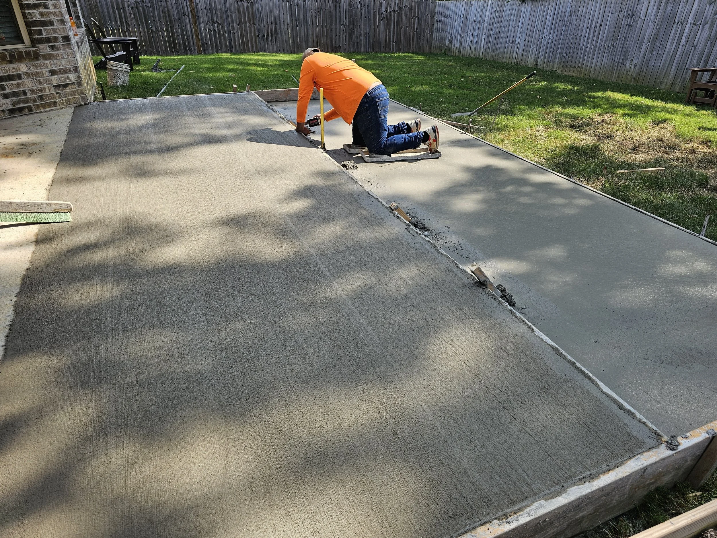 Person kneeling on a concrete patio, working on a small construction or renovation project with tools, next to a lawn and wooden fence.